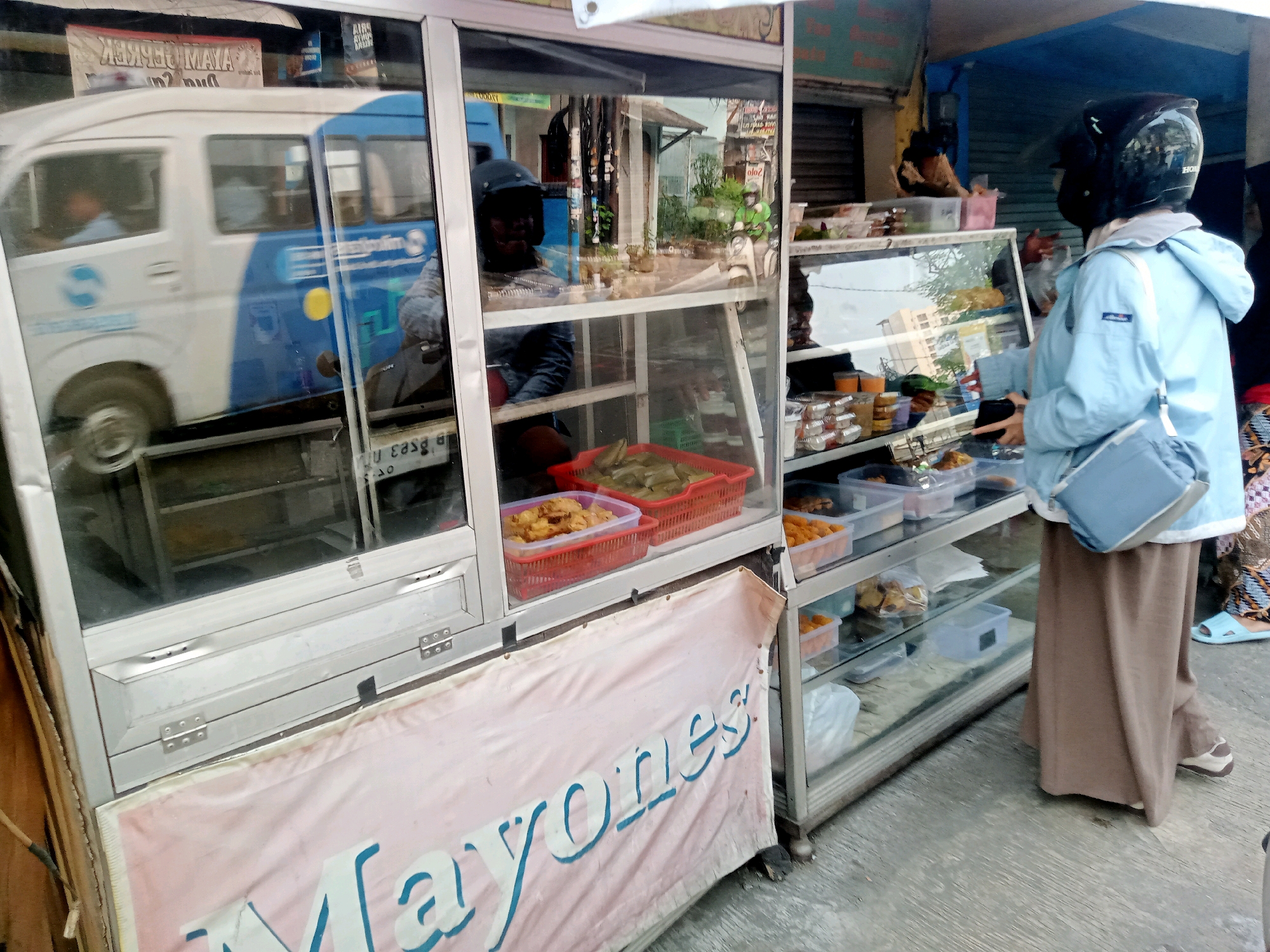 An outdoor food stall is shown in the city of Unknown, Unknown. A vendor wearing a blue jacket, brown skirt, and black helmet is standing in front of a glass display case filled with food. The display case is stocked with various food items in plastic containers, including fried food and wrapped items. A white and blue vehicle, possibly a van, is reflected in the glass of the stall. In the foreground, a pink banner with the word "MAYONES" is partially visible. The ground is a concrete surface. The scene appears to be during the daytime.