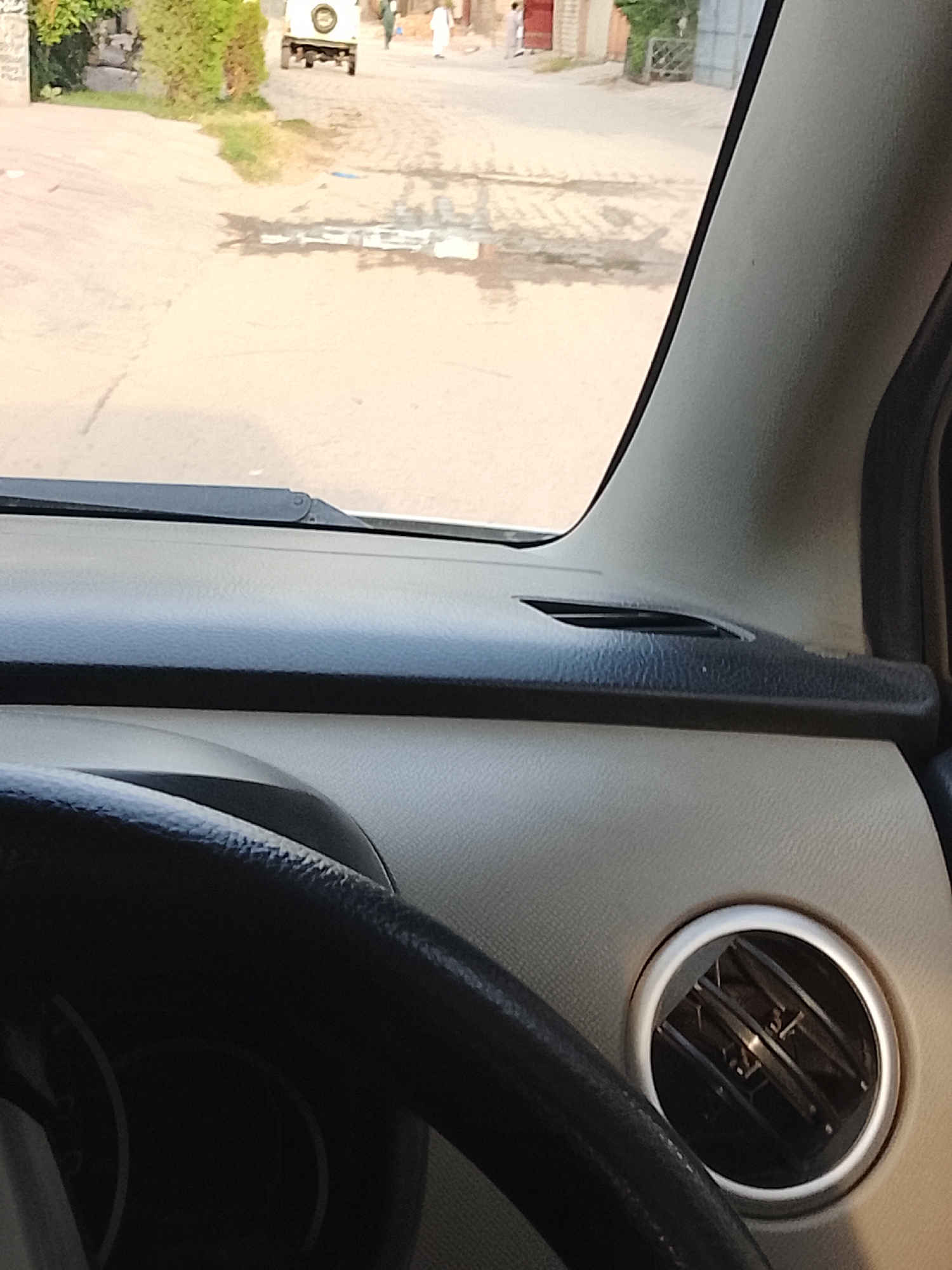 The view from inside a car in Nasheman Colony, Pakistan. The interior of the car, including the steering wheel and dashboard, is visible. Through the windshield, a street scene unfolds. A white vehicle is parked some distance down the road, and two people can be seen walking along the street.