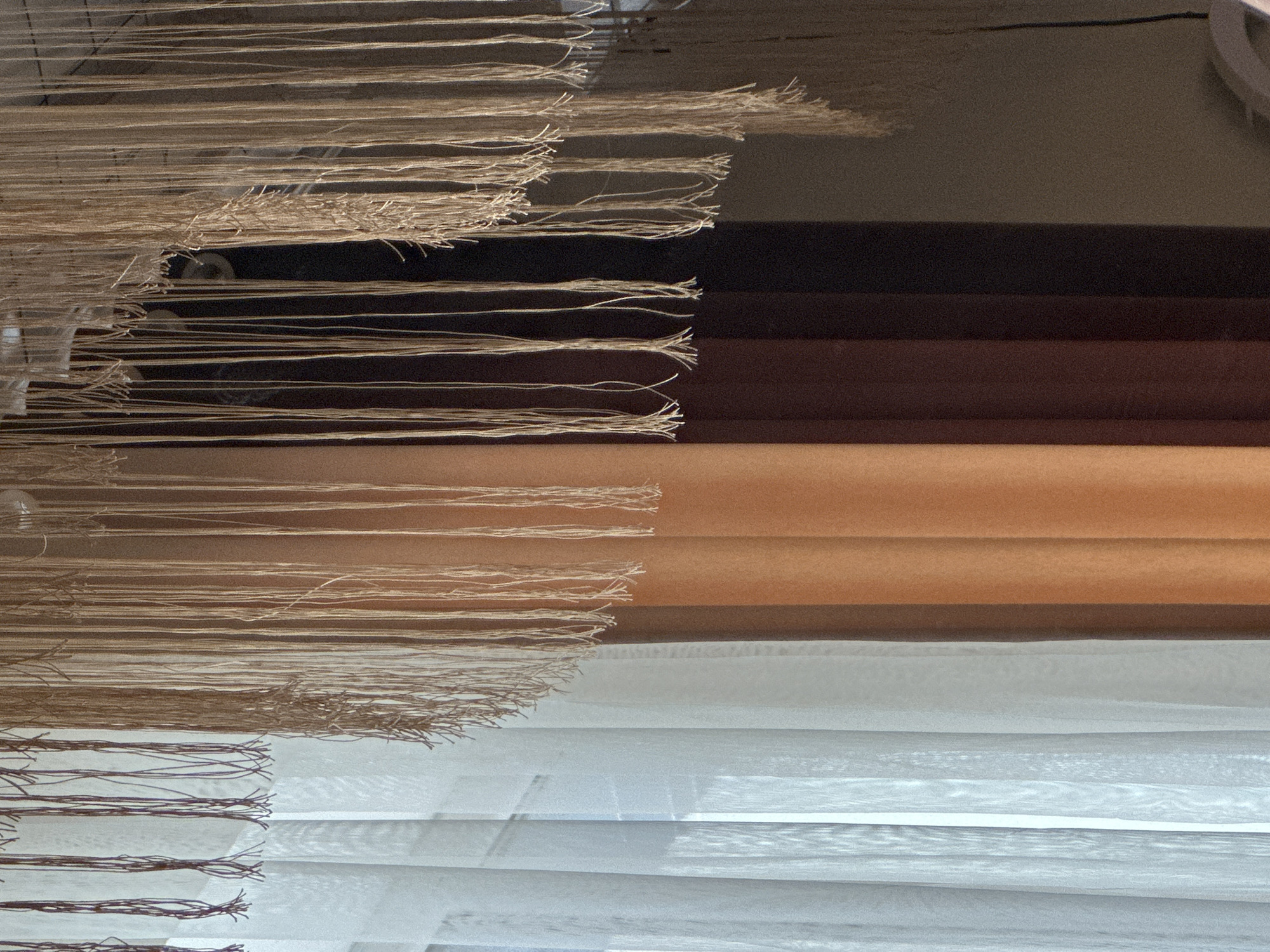 The image depicts a close-up view of a window covered by sheer white curtains on the left, transitioning to vertical blinds in shades of tan, brown, and dark brown. Suspended from above is a decorative element made of numerous hanging strings or cords, varying in thickness and color from light tan to darker brown. Some of these strings are frayed at the bottom, creating a wispy effect. The background is dimly lit and appears to be an interior space. The scene is devoid of any visible people or explicit actions. The location is specified as Kyzyl-Kyya, Kyrgyzstan, suggesting this is an indoor setting within that city.
