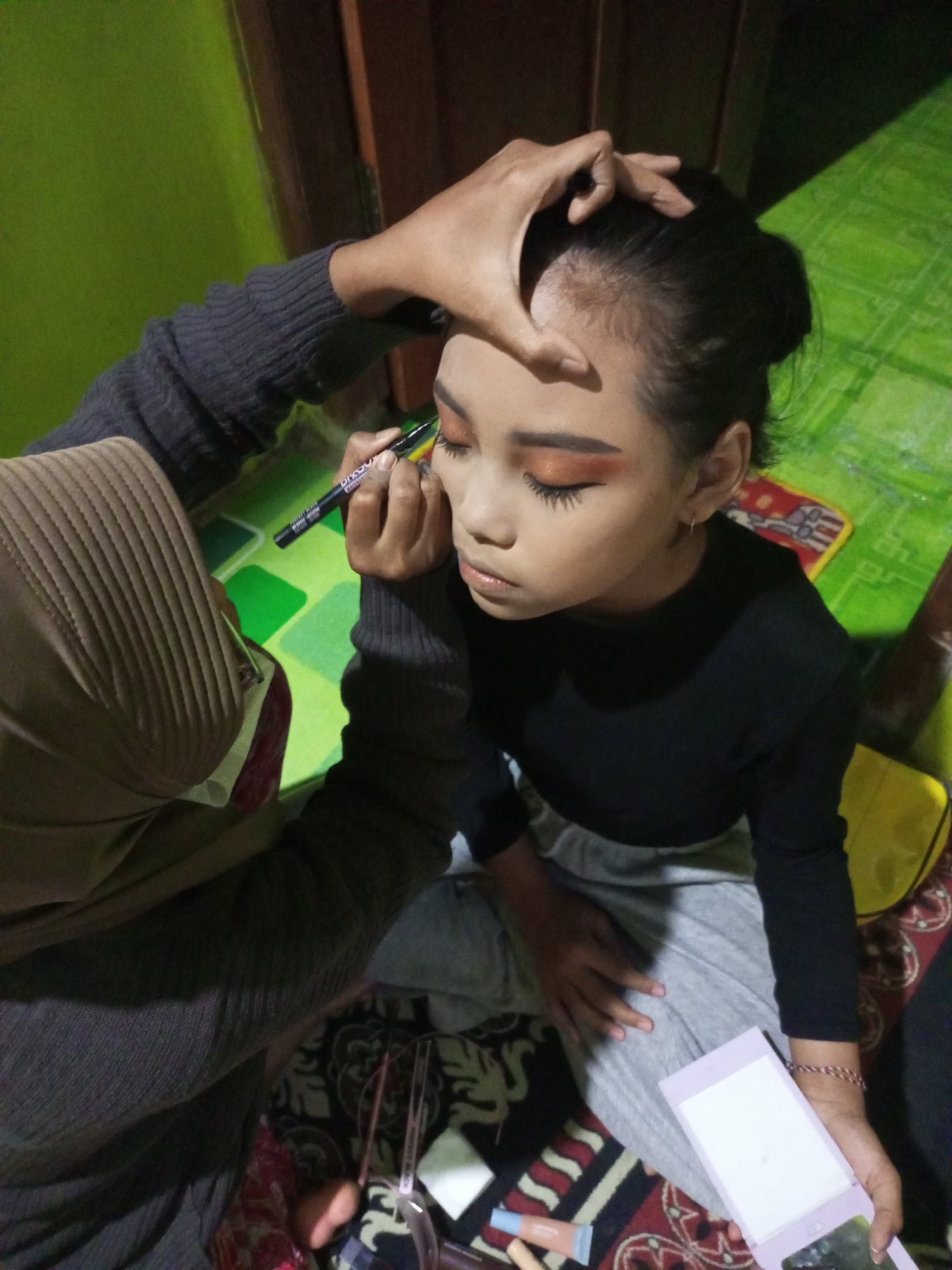 In a room in Nanggulan, Indonesia, a young girl with dark hair styled in a bun is receiving makeup application. Her left hand is raised, and her eyelids are adorned with orange eyeshadow and prominent false eyelashes. An individual, partially visible, is applying eyeliner to the girl's right eye with a black marker. This person is wearing a gray knitted sweater and a brown hijab. The girl is dressed in a black long-sleeved shirt and gray pants. In front of her, a light purple makeup compact rests on a patterned rug. The background shows a green surface, possibly a table, and wooden structures.