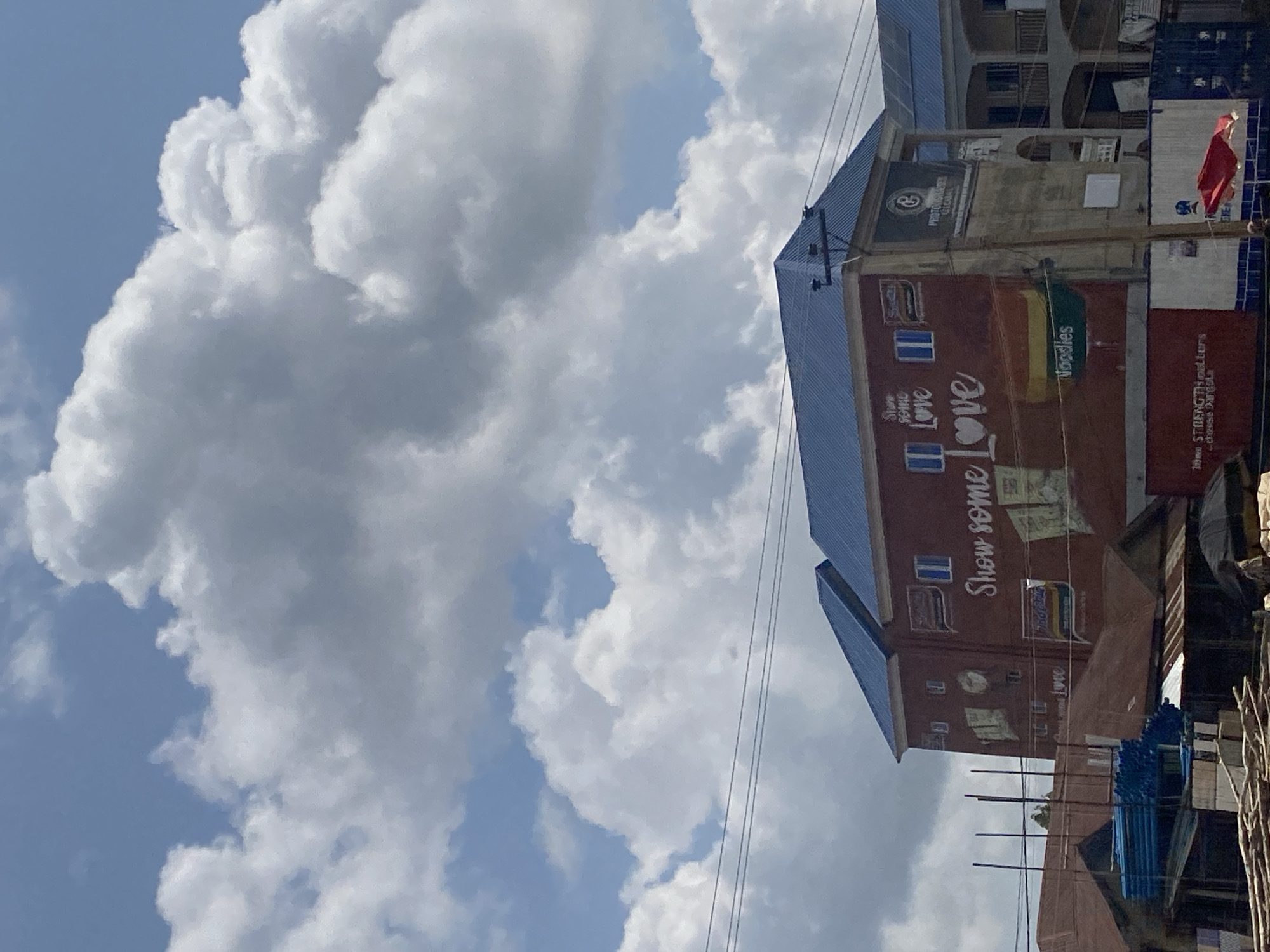 The image depicts a building in Ado, Nigeria, with a prominent mural on its side that reads "Show some love." The building has a blue roof and red walls, with several windows. Various advertisements and signs are visible on the building, including one for "Noodles" and another with a logo that appears to be for "PROTONSTEEL GLOBAL." In the foreground, there are stalls with various goods, including stacks of blue pipes and wooden materials. The sky above is filled with large, white clouds against a blue backdrop. The overall scene suggests a busy commercial or residential area in the city.