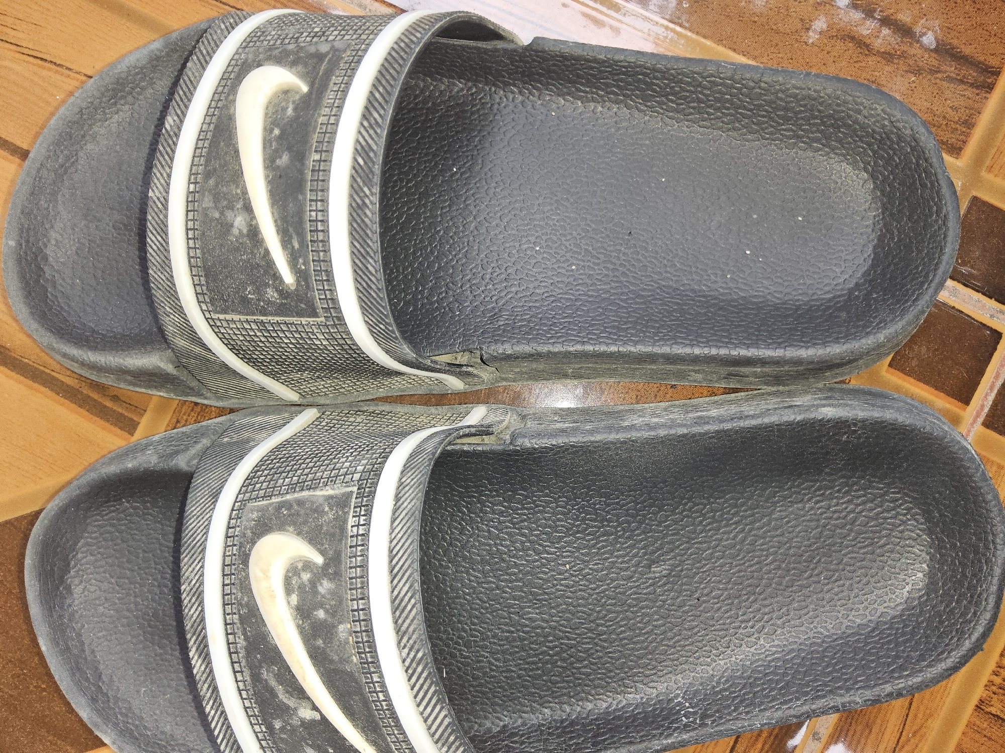 A pair of black Nike slide sandals are positioned side-by-side on a brown, tiled floor. The sandals appear to be worn, with some dust and light smudges visible on the straps, which feature the iconic white Nike swoosh logo. The textured black footbed of each sandal is clearly visible. The background consists of the aforementioned brown tiles, which have a matte finish and appear to be arranged in a grid pattern. The lighting suggests an indoor environment. This image was taken in Payagaboh, Indonesia.