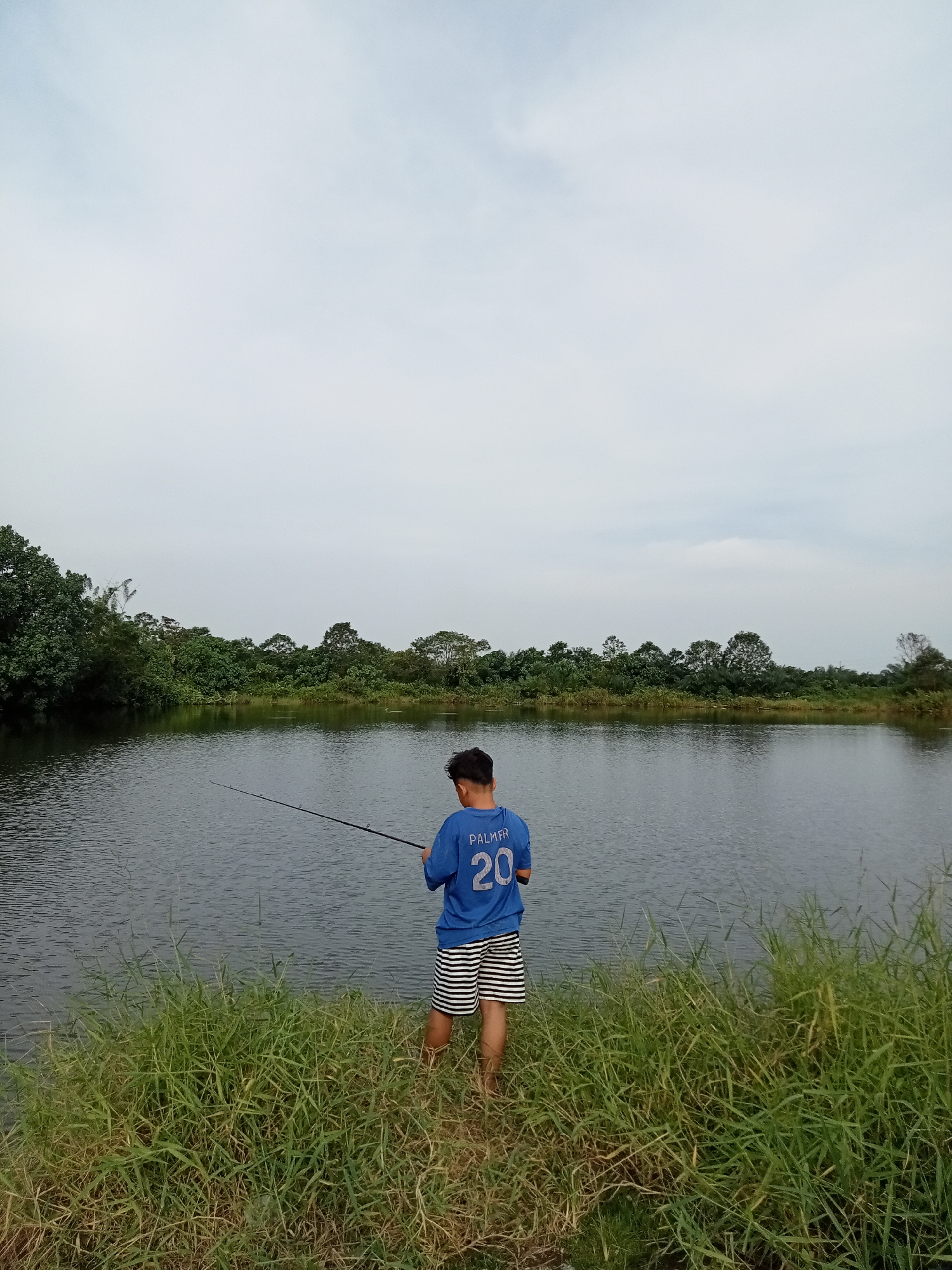 In Kualu, Indonesia, a young male stands facing a calm body of water, holding a fishing rod. He is wearing a blue t-shirt with the name "PALMER" and the number "20" on the back, and black and white striped shorts. He is positioned on the grassy bank of a lake, with lush green trees and vegetation forming a dense treeline in the background. The sky is overcast, with a soft, diffused light. The overall scene depicts a peaceful outdoor activity by the water.