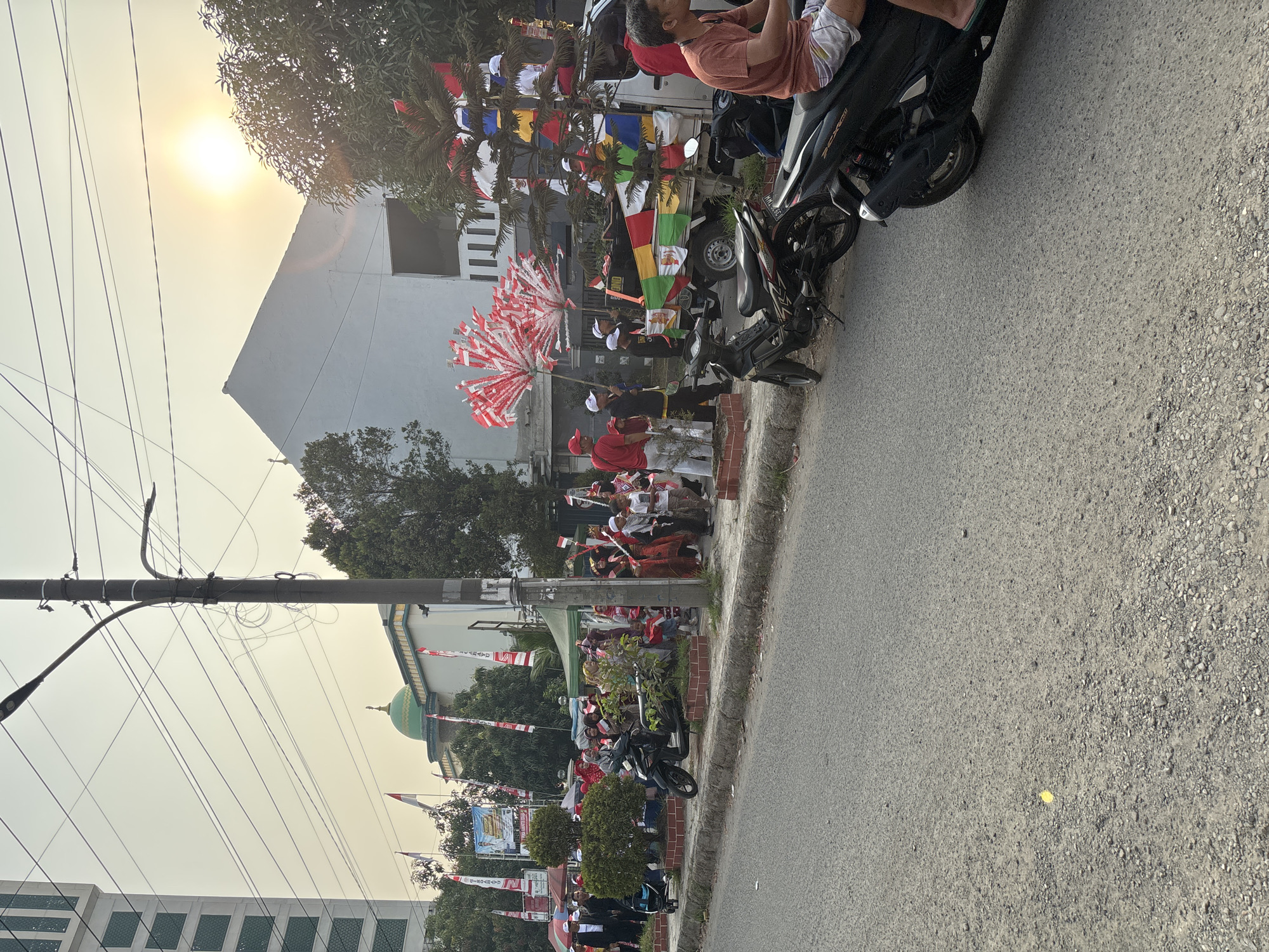 In the city of Telagamurni, Indonesia, a street procession is underway, marked by the presence of numerous Indonesian flags and people dressed in red and white attire. The scene takes place on a street with a grey asphalt surface. On the left side of the street, a tall, modern building with green windows stands prominently. Adjacent to it, a mosque with a green dome and white walls is visible. A power pole with multiple wires runs vertically through the image, positioned between the building and the procession.
The procession includes a group of people walking, many holding small red and white flags or larger, fan-like decorations made of red and white flags. Some individuals are dressed in traditional Indonesian clothing, including batik. A black motorcycle is parked on the sidewalk, and further down the street, another black motorcycle is visible, with a person on it wearing a red shirt and checkered shorts.
In the background, a small truck is decorated with colorful flags, including Indonesian flags. The sky is hazy with the sun shining brightly, creating a glare. Trees with green foliage are scattered throughout the scene. A tent is set up on the sidewalk, and banners with images of people are displayed. The overall atmosphere appears festive, suggesting a celebration or parade.