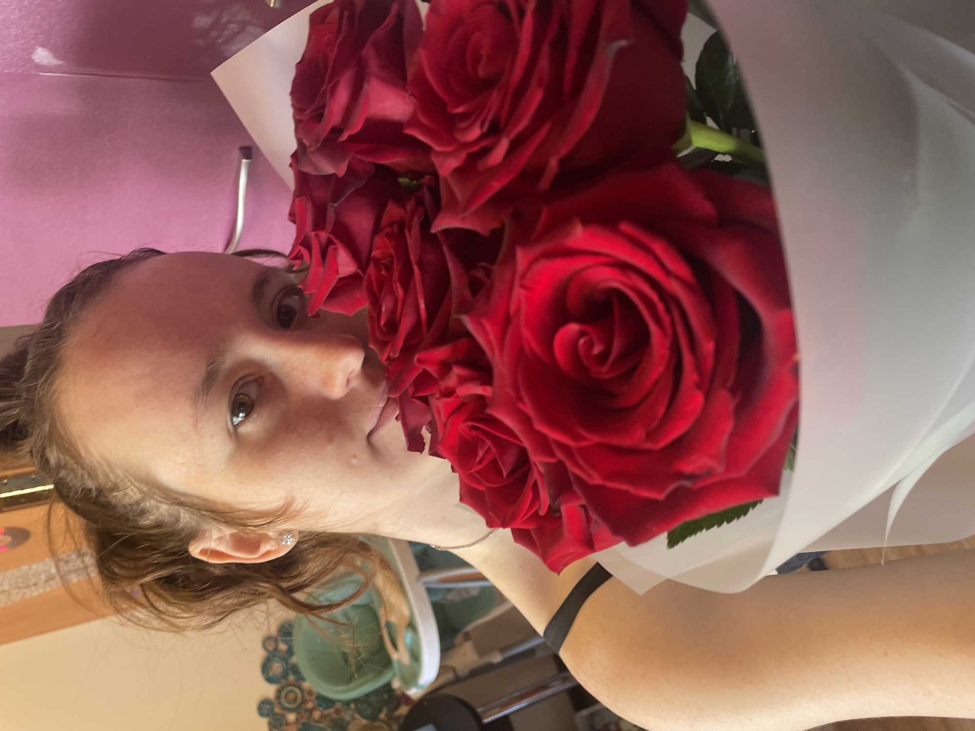 A young woman is captured in a close-up, holding a large bouquet of dark red roses. Her face, seen in profile on the left side of the frame, shows a pleasant expression with a slight smile, and her left eye is visible. She has light skin and brown hair, which appears to be styled back, revealing a small, sparkling earring in her left ear. A thin silver necklace is also visible around her neck.
The bouquet of deep red roses dominates the right side of the image, filling a significant portion of the foreground. The roses are tightly clustered, showcasing their rich color and intricate petals. They are wrapped in a soft, light-colored material, likely white or cream paper or plastic, which gently folds around the blooms. Some green foliage is visible peeking from between the roses and the wrapping.
The background is softly blurred, suggesting an indoor setting. Behind the woman's head, there is a plain pink or magenta wall or surface. To her lower left, a warm-toned surface, possibly wood, features a decorative circular pattern. Part of a light green object, possibly a bowl or planter, is also vaguely visible in the soft-focus background. The overall scene conveys a feeling of contentment and appreciation for the flowers. There is no visible text in the image.