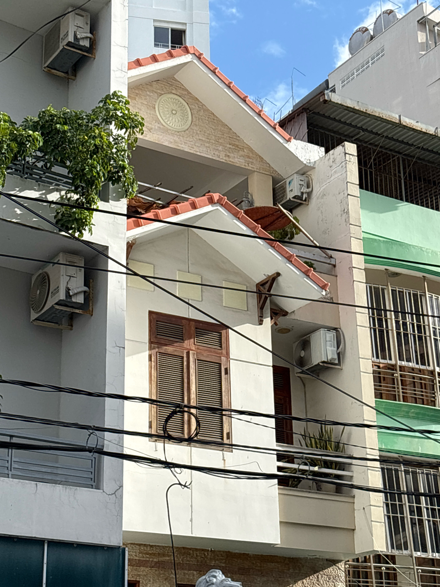 The image shows a view of buildings in the city of Nha Trang, Vietnam. The scene is dominated by a white building with a red-tiled roof, featuring a prominent window with wooden shutters. An air conditioning unit is mounted on the exterior wall of this building. To the left, another white building is visible, also with an air conditioning unit attached. Overhead, a network of black electrical wires crisscrosses the frame, obscuring parts of the buildings. Behind the main white building, a section of a green-colored building with barred windows can be seen. The sky above is a clear blue. The overall impression is of a dense urban environment with a focus on residential architecture and utility infrastructure.