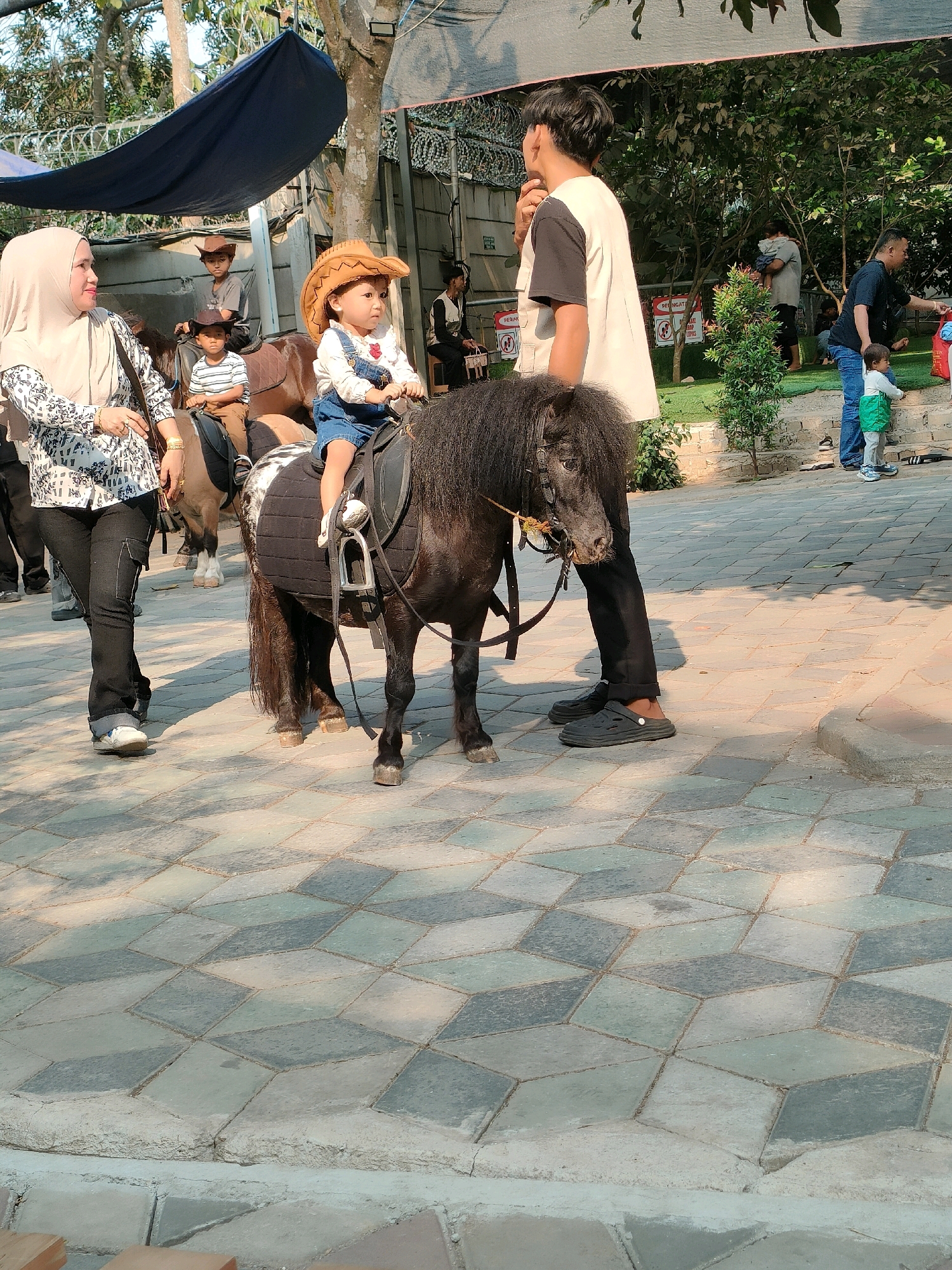 In Cileduk, Indonesia, a group of people are gathered at what appears to be an outdoor amusement or recreational area. The focus is on a pony ride activity. A young child, dressed in a denim outfit and a brown cowboy hat, is seated on a small black pony, holding the reins. To the left, a woman wearing a light-colored hijab and a patterned shirt is walking alongside the pony, appearing to be supervising. Behind her, another child is also seated on a pony, wearing a cowboy hat.
In the background, several other individuals are visible. A young man in a white shirt with black sleeves stands to the right of the child on the pony, seemingly guiding it. Further back, adults and children are scattered around the area. The environment includes trees and a paved pathway with a geometric pattern of square and diamond-shaped tiles. A dark blue tarp is visible overhead, providing shade. The scene suggests a family-friendly outing.