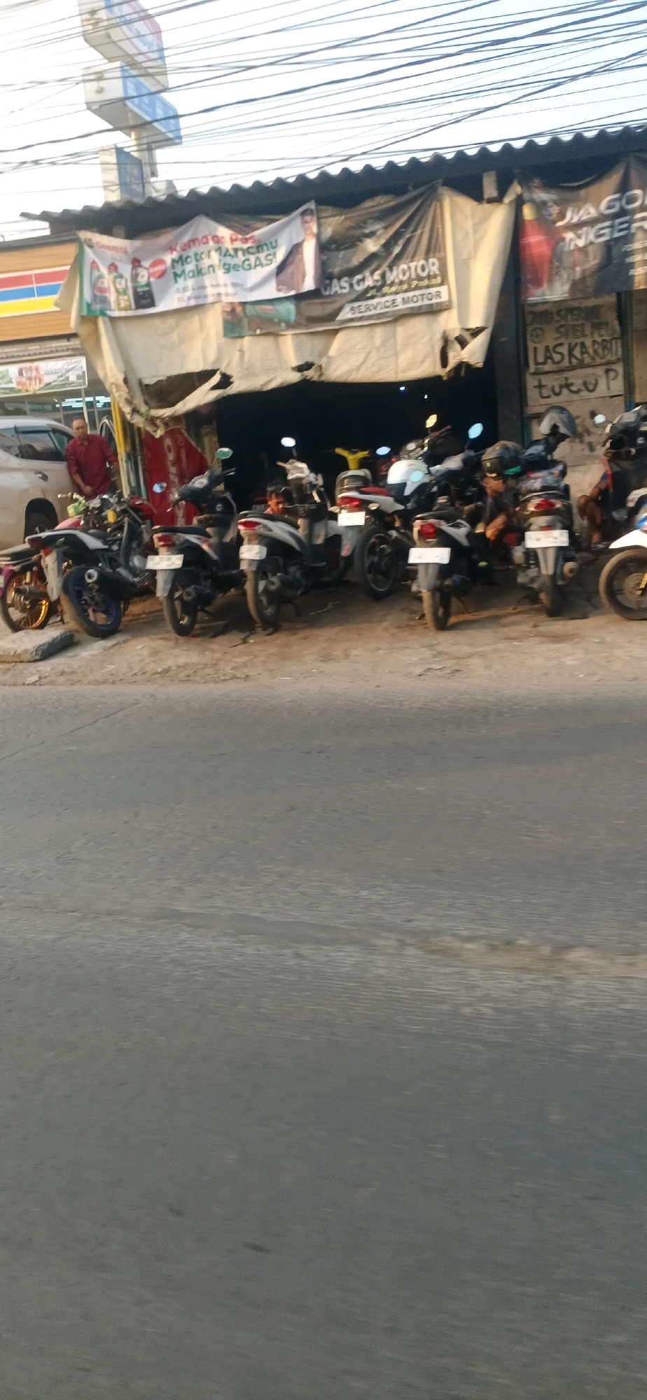 The image depicts a motorcycle repair shop, likely located in Surabaya, Indonesia, given the Indonesian text on the banners. The shop is open, with several motorbikes parked outside. A man in a red shirt stands by a white car on the left, while another person, possibly a mechanic, is seen kneeling amongst the motorbikes inside the shop. Banners advertising "GAS GAS MOTOR" and "SERVICE MOTOR" are hung above the shop. The scene is set against a backdrop of overhead power lines and a bright sky, suggesting it is daytime. The ground is a rough, paved surface, and the overall impression is of a busy, functional repair shop.