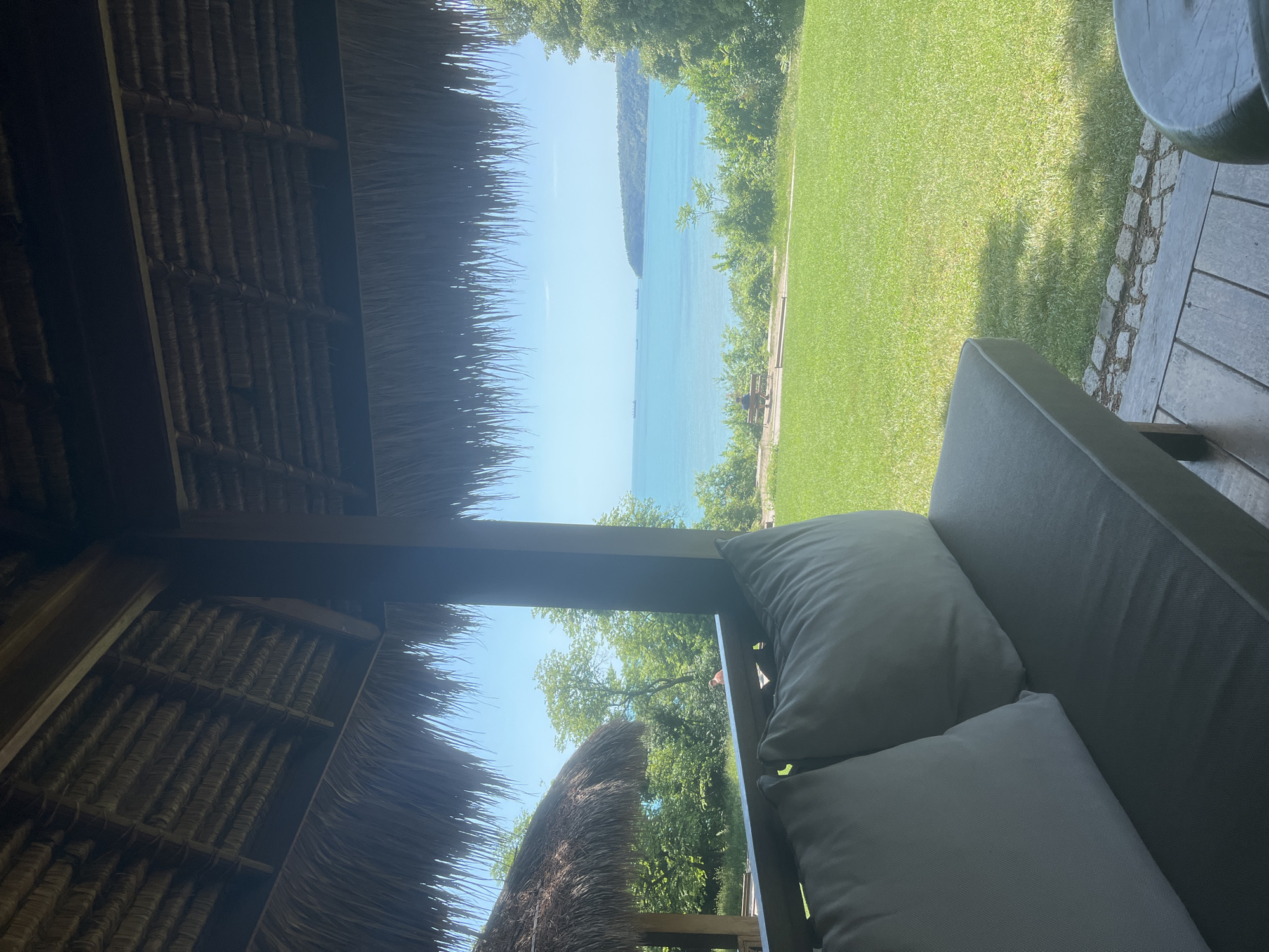 This image captures a tranquil outdoor scene, viewed from the shade of a thatched-roof structure, likely a patio or veranda, overlooking a scenic coastal landscape in Varna, Bulgaria.
In the immediate foreground, the rustic texture of a traditional thatched roof is visible on the left side, providing a sense of natural shelter. On the right, a comfortable outdoor daybed or sofa, upholstered in dark grey fabric and featuring two light grey cushions, invites relaxation. Adjacent to this seating, a segment of a wooden deck is visible, bordered by a strip of rough stones or pebbles.
Beyond the shaded area, a vibrant green lawn stretches across the midground, bathed in bright sunlight. Distinct shadows are cast upon the grass, suggesting the presence of unseen trees overhead. The lawn seamlessly transitions into a calm, expansive body of clear, turquoise water, characteristic of a sea or large bay. Several small boats or ships can be seen gently floating in the distance on the water. A verdant, tree-covered coastline or distant hills define the far horizon across the water.
The sky above is a brilliant, cloudless blue, indicating a sunny and clear day. The bright natural light and visible shadows suggest it is daytime, possibly mid-morning or afternoon. The overall atmosphere conveyed is one of peace, relaxation, and natural beauty. No people are visible in the frame, and there is no discernible text.