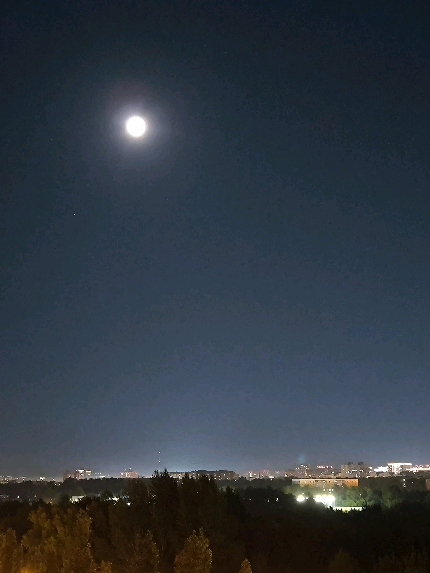 A full moon shines brightly in the dark night sky above a city skyline. The moon is located in the upper left quadrant of the image. Below the moon, a small, bright star or planet is visible. The lower portion of the image shows a distant cityscape with buildings illuminated by artificial lights, nestled among dark trees. The scene appears to be overlooking a residential area or possibly a parkland within the city. The image captures a serene night view of an urban environment under a luminous moon. The location is the city of Kazan, Russia.