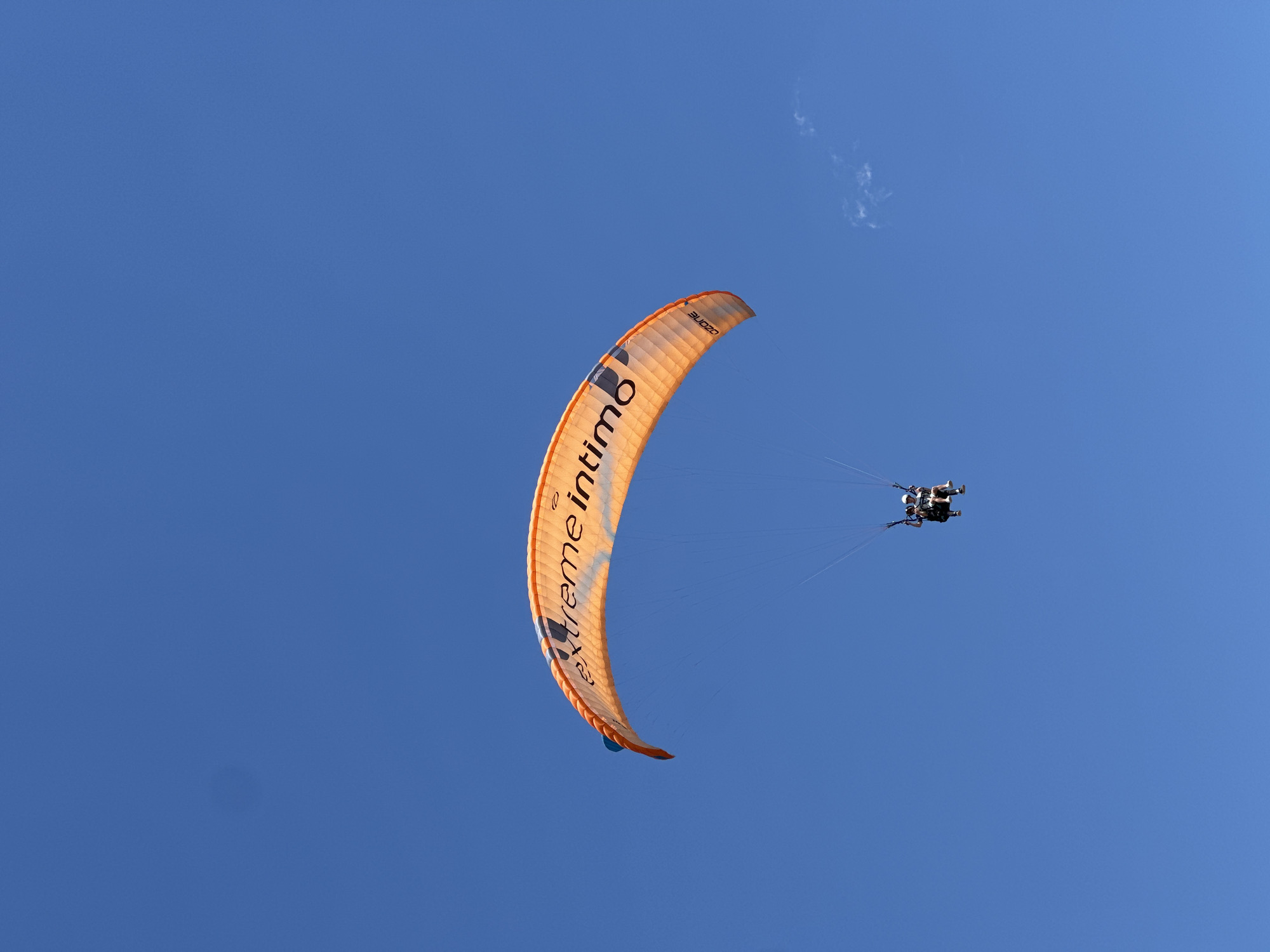 An orange paraglider with the words "EXTREME INTIMO" and "OZONE" printed on it is soaring through a clear blue sky. Two people are seated in the harness below the paraglider. The paraglider is positioned in the upper center of the frame, angled slightly to the right. The two individuals are visible in the lower center of the image. The sky is uninterrupted except for a small wisp of cloud on the right side of the frame. There is no indication of the city of Boreti, Montenegro in the image.