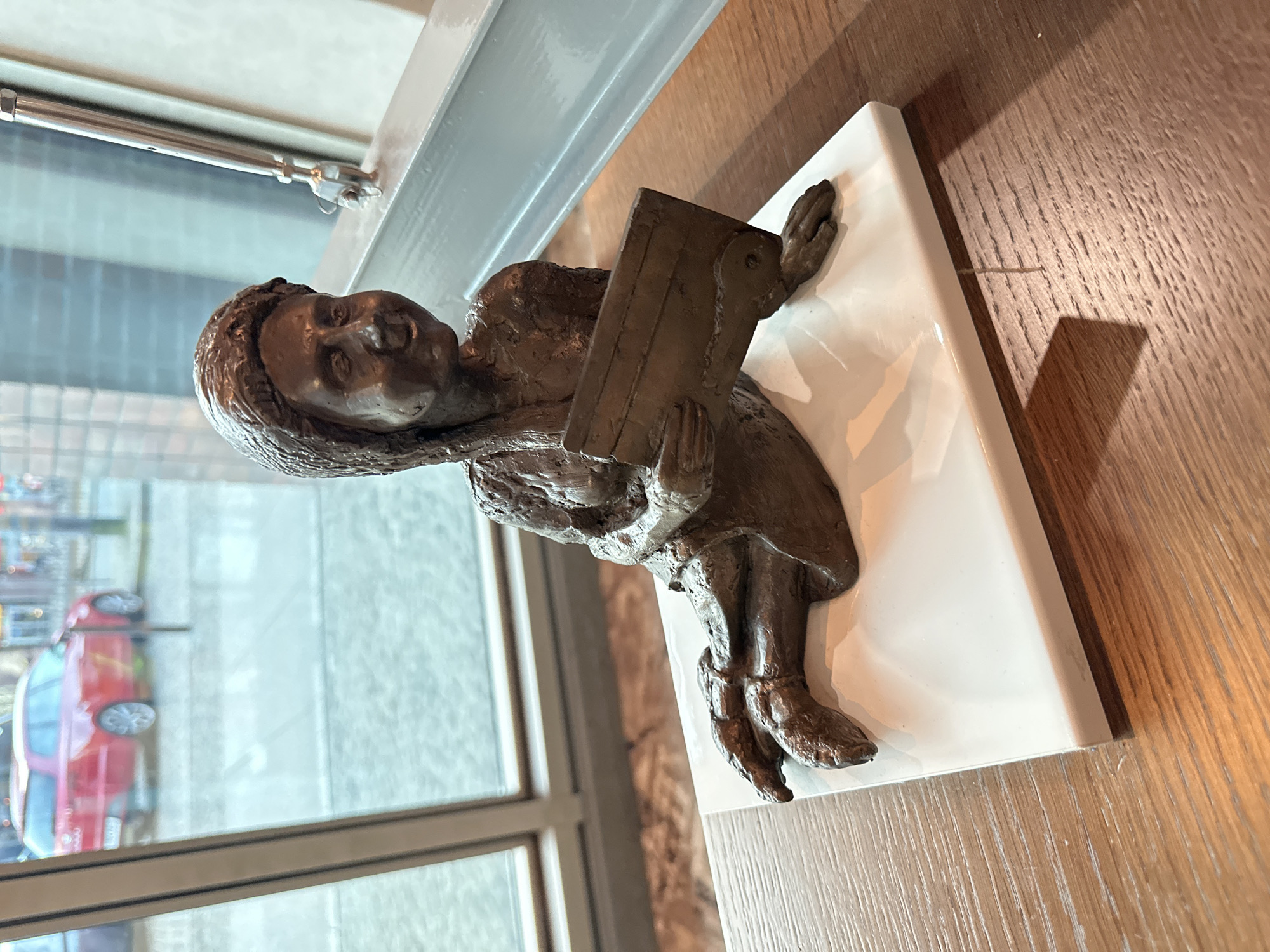 A bronze sculpture of a child reading a book sits on a white base on a wooden surface, possibly a table. The child is wearing a head covering and a dress, and the sculpture is positioned near a window. Outside the window, a red car is visible on the street. This sculpture is part of the "Wrocław Dwarfs" project in Wrocław, Poland.