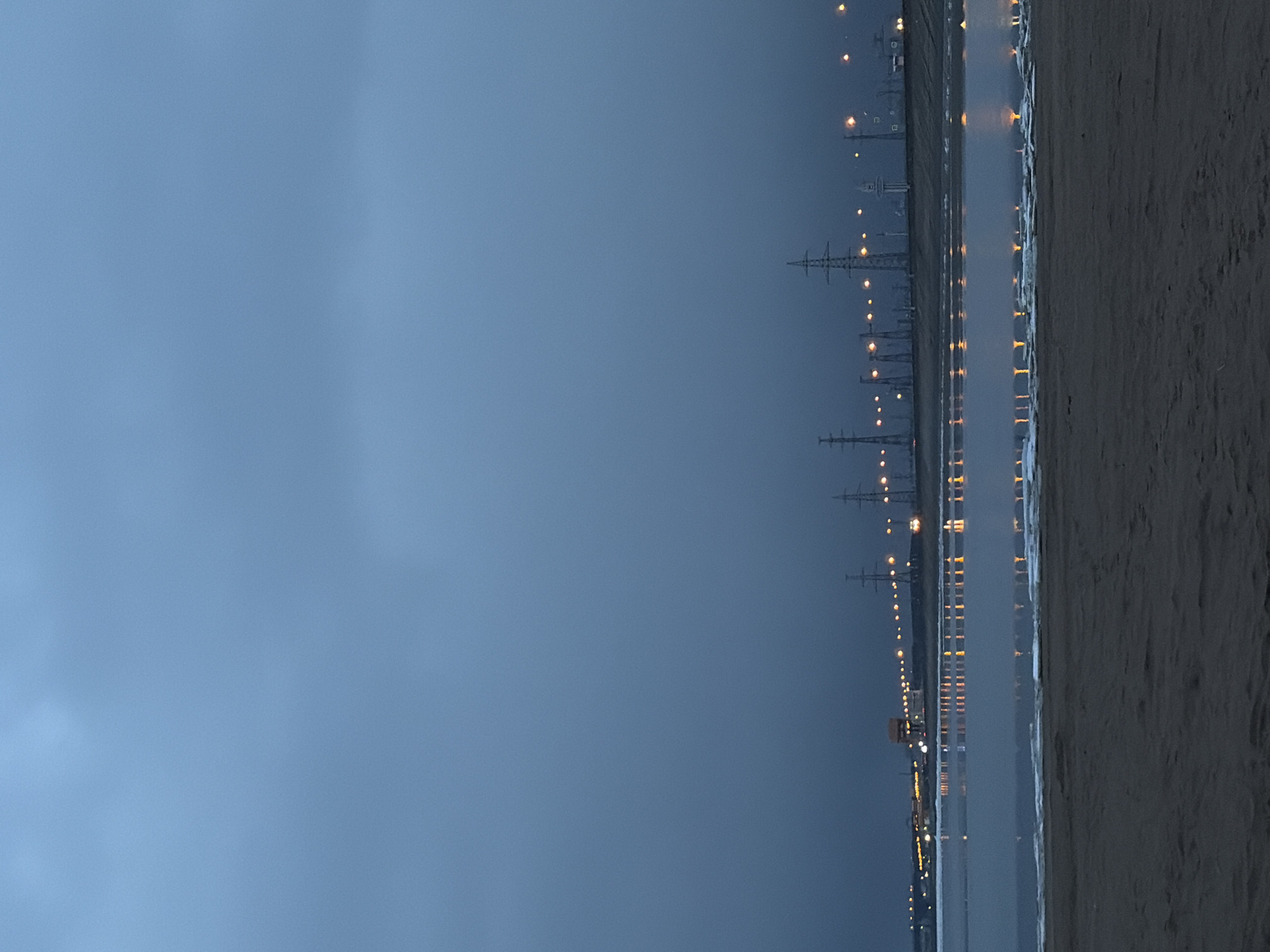 The image captures an industrial waterscape at twilight or night, with the location specified as Noviy, Russia. The scene is characterized by a muted color palette dominated by blues, greys, and dark browns, accented by bright artificial lights.
In the foreground, a dark, textured expanse, likely a sandy or rocky shoreline or beach, occupies the right side of the frame. This area appears largely desolate and devoid of any human presence or activity.
To the left of the shore, a calm body of water stretches into the distance. This water reflects a series of bright, warm-toned lights from the opposite bank, creating shimmering vertical streaks on the dark surface. These reflections suggest a still body of water, possibly a large river, lake, or coastline.
Along the far bank of the water, a long line of prominent artificial lights is visible, illuminating what appears to be a road, industrial facility, or distant urban development. Behind these lights and extending further into the background, numerous tall electrical transmission towers or pylons stand silhouetted against the dark sky, reinforcing the industrial character of the location.
The sky, filling the upper left portion of the image, is a deep, overcast blue-grey, suggesting either heavy cloud cover or the profound darkness of night. The overall impression is one of solitude and a stark, industrial environment. No people, animals, or specific events are depicted, and there is no visible text.