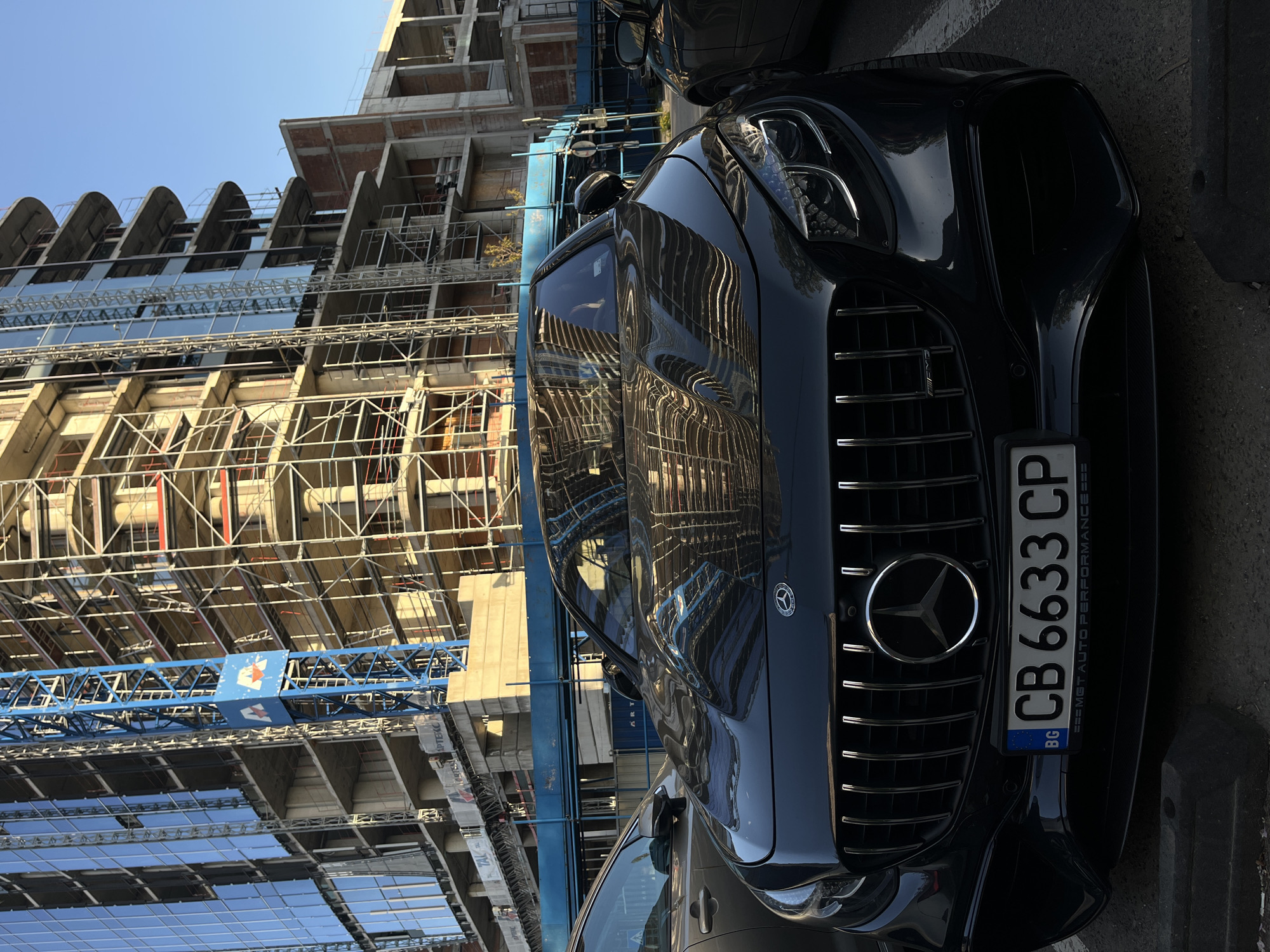 The image captures a static scene in an urban environment, featuring a luxury car parked near a large construction site on a clear, sunny day. The perspective is tilted, showing the subjects from an elevated, slightly rotated angle.
The primary subject in the foreground is a dark-colored Mercedes-Benz car, identifiable by its prominent three-pointed star emblem on the grille. The car has a sleek, polished finish that highly reflects its surroundings, showcasing distorted reflections of the construction site and the blue sky. The front of the car, including its grille and headlights, is clearly visible.
Behind the car, a multi-story building under construction dominates the background. The building features exposed concrete structures, numerous support beams, scaffolding, and partially installed glass facades on its lower and mid-levels. Distinct arched concrete elements are visible on one side of the structure. A large blue construction crane, with a lattice boom, extends across the middle ground of the scene, indicating ongoing building work.
Notable details include:
- The bright, clear blue sky, suggesting good weather conditions.
- The intricate reflections on the car's body, which provide a distorted mirror image of the construction work.
- Visible text on the car's license plate reads "BG CB 6633 CP", indicating the car is registered in Bulgaria. Below the license plate, the text "=== MGT AUTO PERFORMANCE ===" is partially visible.
- On the blue crane, a logo with an abstract 'E' shape in white on a blue background is discernible. Additional text, "APTEKC" and "TAL", can be seen on vertical banners near the construction site.
The image portrays a blend of modern urban development (the construction) and luxury (the car), set against a backdrop of everyday city life.