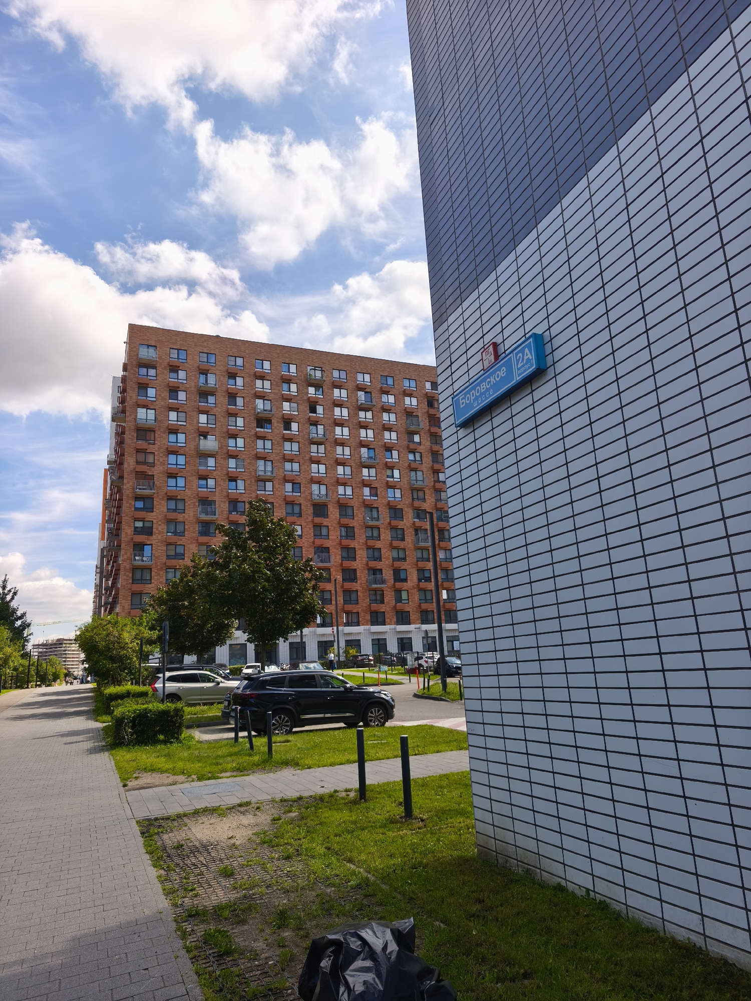 The image depicts a street scene in Moscow, Russia, specifically featuring modern residential buildings and a street sign. In the foreground, a grey tiled sidewalk runs alongside a patch of green grass. A black trash bag is visible on the grass to the right. Further back, a dark SUV is parked on the side of the road, with a light-colored SUV parked behind it.
Dominating the mid-ground is a tall, brick-faced apartment building with numerous windows and balconies. To its left, a lighter-colored, modern building with a blue and white tiled facade is prominent. A blue street sign is attached to this building, reading "Боровское шоссе" (Borovskoye Shosse) and "2А корпус 1" (2A Building 1). The sky is blue with scattered white clouds. In the distance, construction cranes are visible, suggesting ongoing development. No people are clearly identifiable or engaged in any specific actions. The overall environment appears to be a well-maintained urban residential area on a clear day.