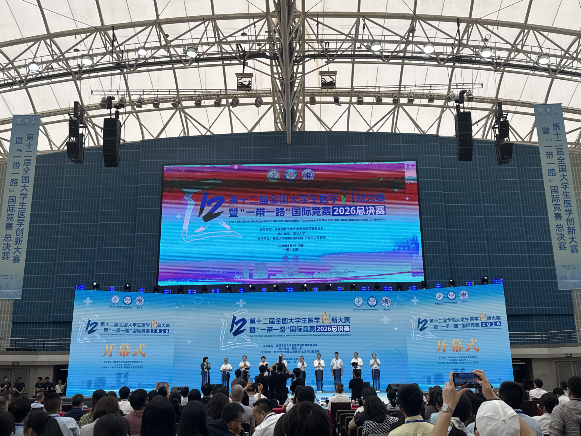 The image captures a large indoor event, likely an opening ceremony, at the University of Fudan's Jiangwan Campus in Yangpu District, China. A prominent LED screen displays event details in Chinese and English, announcing "The 12th China Undergraduate Medical Innovation Tournament & The Belt and Road International Competition 2026 Finals". The screen also shows the event title as "Opening Ceremony".
A group of individuals, presumably participants and organizers, stand on a stage in front of the screen, some holding microphones or gesturing. Spectators are seated in rows of chairs, facing the stage and screen, with many holding up smartphones to record the event. The environment is a large, modern hall with a high, arched ceiling structure supported by metal beams. Large black speakers are suspended from the ceiling. Banners with Chinese text are visible on the left and right sides of the stage, further identifying the event. The overall atmosphere appears to be formal and celebratory.