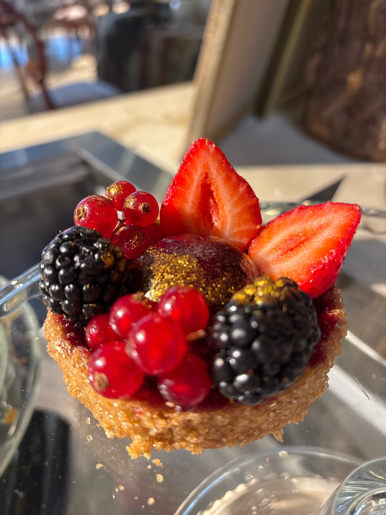 A close-up, eye-level shot captures a vibrant, miniature tart adorned with fresh berries and edible gold glitter. The tart's crust appears golden brown and slightly granular. It is topped with two halves of a bright red strawberry, a cluster of small, red currants, and two dark, plump blackberries. A small, dark, glossy sphere, possibly a fruit gel or a chocolate dome, is positioned in the center of the tart, dusted with shimmering gold particles. The background is softly blurred, showing reflections of what appears to be an indoor environment with some daylight. The overall impression is one of a meticulously crafted dessert, likely served in a cafe or patisserie. The location is not specified in the image.