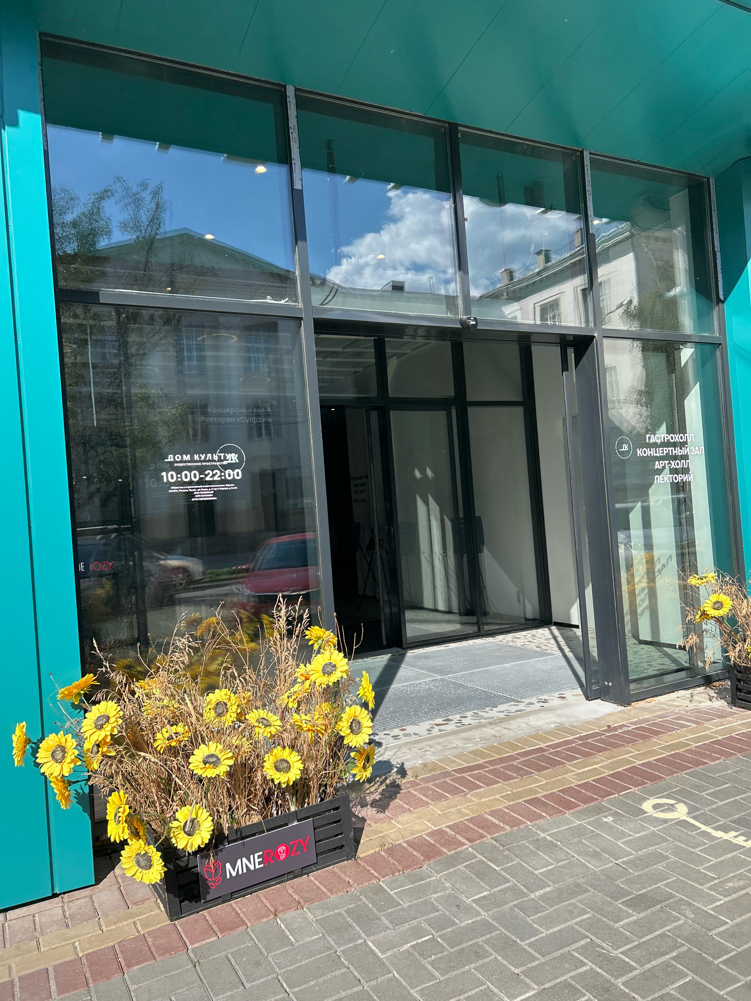 The image shows the entrance of the "Dom Kultury" (House of Culture) in Tomsk, Russia. The building features a modern facade with large glass windows and a teal-colored overhang. In front of the entrance, a planter box is filled with artificial sunflowers and dry foliage, with a black sign that reads "MNEROZY".
Visible on the glass door is signage indicating "ДОМ КУЛЬТУРЫ" (House of Culture) and operating hours from "10:00-22:00". Another sign on the right side of the entrance lists various areas within the complex: "ГАСТРОХОЛЛ" (Gastro Hall), "КОНЦЕРТНЫЙ ЗАЛ" (Concert Hall), "АРТ-ХОЛЛ" (Art Hall), and "ЛЕКТОРИИ" (Lectures).
The entrance itself has automatic sliding glass doors. The ground in front of the entrance is paved with interlocking bricks in a herringbone pattern, with a yellow directional arrow painted on the grey section of the paving. Reflections in the glass show buildings and the sky with clouds. Two cars are visible parked on the street beyond the entrance, one red and one silver. The overall impression is of a public cultural center on a sunny day.