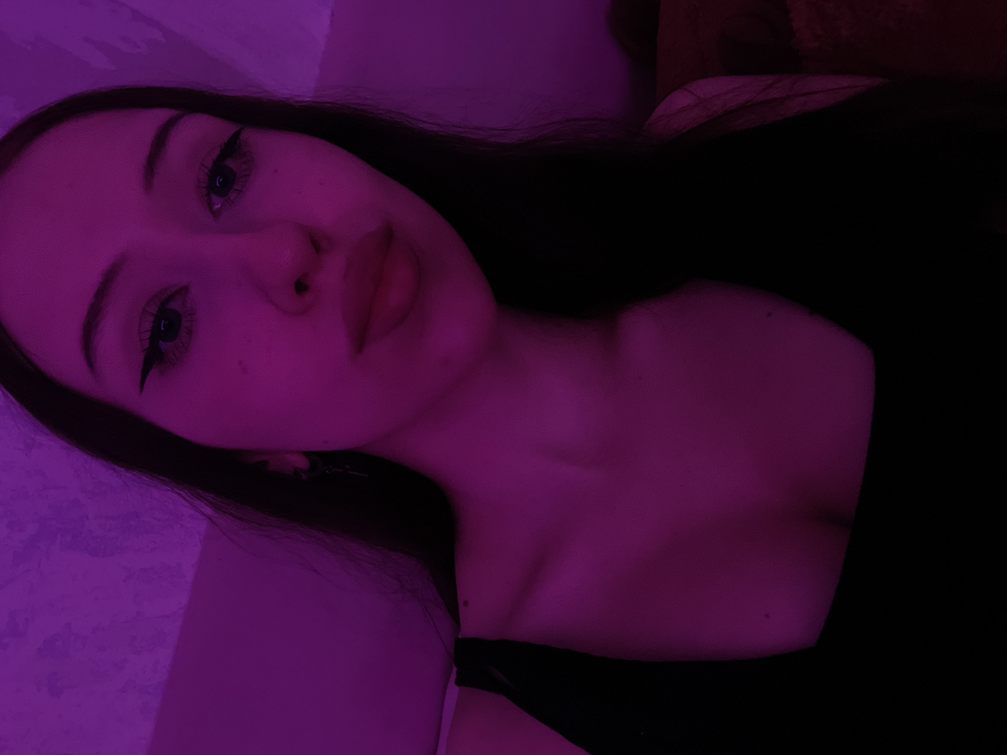 The image is a close-up, indoor selfie of a young woman illuminated by purple lighting. She has dark hair, prominent eyeliner, and full lips. She is wearing a black top. The background appears to be a wall with a textured surface, also lit in purple. No specific location in Votkinsk, Russia can be identified from the image.