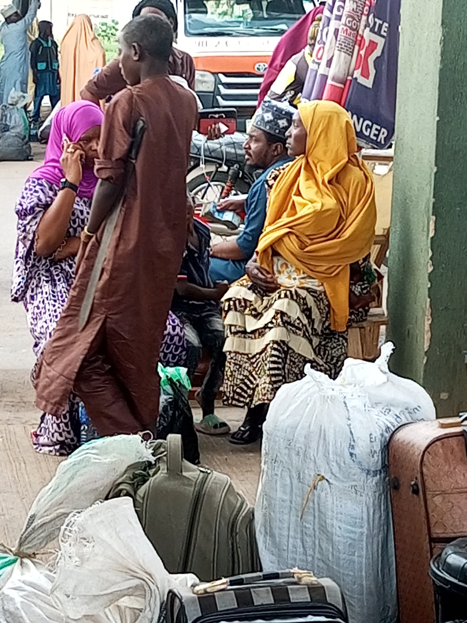 The image shows a group of people with luggage, likely at a transportation hub, with a building and a vehicle in the background. The setting appears to be outdoors, possibly on a street or a waiting area.
In the foreground, several pieces of luggage are visible, including large white plastic bags, a brown suitcase, and a dark green duffel bag. A person in the center is wearing a long brown garment and appears to be interacting with another person on the left, who is wearing a purple headscarf and a patterned dress, and is holding a phone to her ear. To the right of the person in brown, a woman in a bright yellow headscarf is seated, wearing a patterned skirt. Another man is visible behind the woman in yellow, wearing a blue shirt and a patterned cap. In the background, there are other people standing and walking, as well as parts of a vehicle and some colorful banners or fabric.
The overall impression is one of travel or waiting for transport, with a focus on the people and their belongings. The presence of multiple individuals and luggage suggests a public space where people are on the move. The style of dress and the environment hint at a location in Nigeria, possibly Gombe city, given the context provided, where such scenes are common.