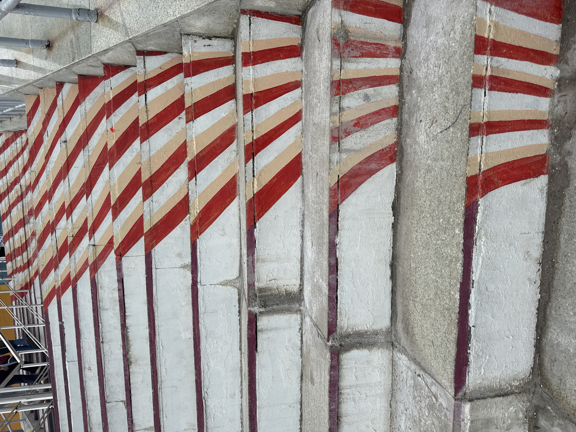 The image shows an outdoor concrete staircase with red, white, and beige diagonal stripes painted on the risers and some of the treads. The paint is chipped and worn in places, suggesting age and exposure to the elements. The stairs are wet in some areas, possibly from recent rain. In the background, there are metal railings and the legs of several people are visible, dressed in casual clothing. The setting appears to be a public space in Ufa, Russia, likely an elevated walkway or an entrance to a building or metro station.