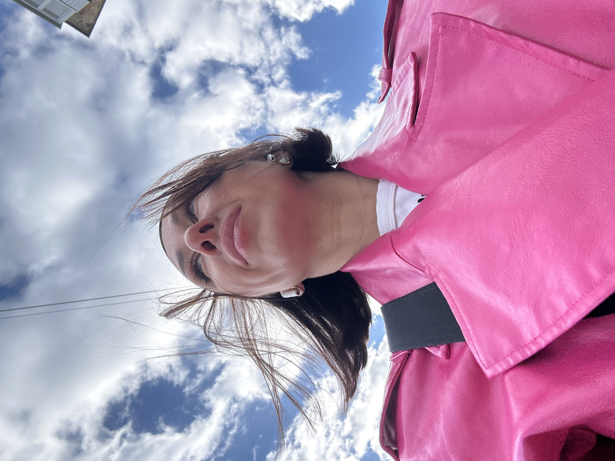 A low-angle shot depicts a woman wearing a pink leather jacket against a backdrop of a blue sky dotted with clouds. She is slightly angled towards the camera, revealing her face, which includes hair pulled back, an earring, and what appears to be an earbud. Power lines are partially visible against the sky. The location of Votkinsk, Russia, cannot be determined from the image.