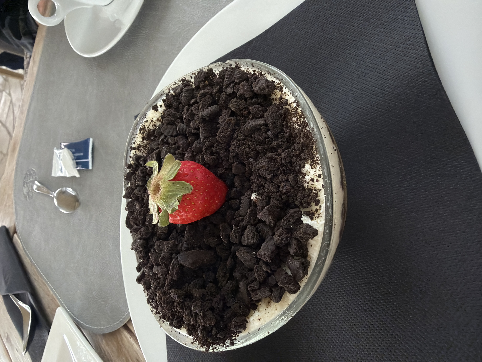 The image depicts a dessert served in a glass bowl, placed on a table. The dessert appears to be a creamy base topped with crushed chocolate cookies, and a single strawberry is placed in the center. Nearby are a white saucer with a cup, a small spoon, and a sugar packet. The table is covered with a light gray placemat on one side, and a black textured surface on the other. Given the presence of a white plate and cutlery, it can be inferred that this is a restaurant or cafe setting. The image does not include sufficient detail to conclusively ascertain the location as Budva, Montenegro.