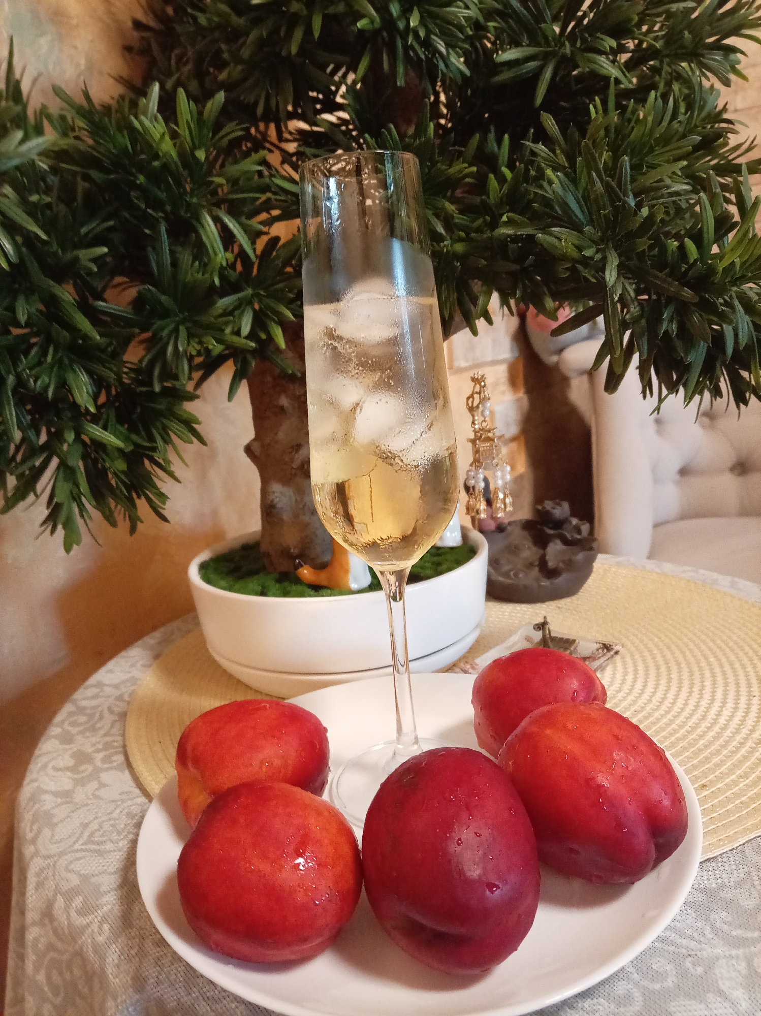 In the city of Yessentuki, Russia, a tabletop scene is captured, bathed in natural light. A white plate, adorned with a delicate pattern, holds five ripe nectarines, their skins glistening with water droplets. To the left of the plate, a tall, slender champagne flute is filled with a pale yellow, effervescent liquid and ice cubes. The glass stands amidst a lush, green artificial plant, whose branches cascade over the scene. In the background, a small, white pot contains a mossy base, hinting at a decorative display. A glimpse of ornate decor and a plush armchair can be seen further back, suggesting a domestic setting.