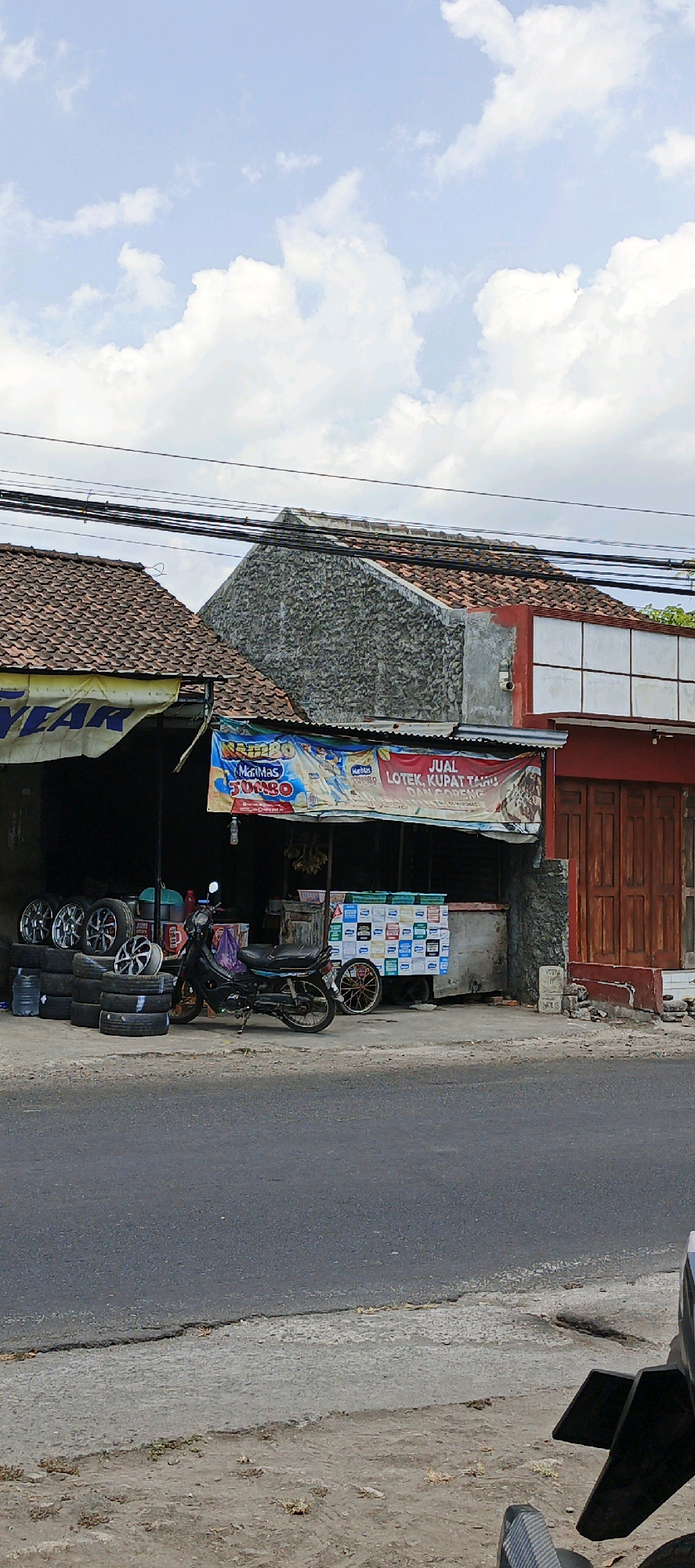 The image captures a street scene in Godean, Indonesia, with a focus on a small roadside establishment. The establishment appears to be a shop or food stall with a covered area. In front of it, several car tires are stacked, and a motorcycle is parked. There is also a food cart decorated with various colorful panels. A banner hangs above the stall, displaying text and images, including what appears to be food items and branding. Behind the stall, buildings with tiled roofs are visible, some constructed with textured stone. The sky above is partly cloudy. The area in front of the establishment consists of a dirt and gravel surface, which transitions into a paved road in the foreground.