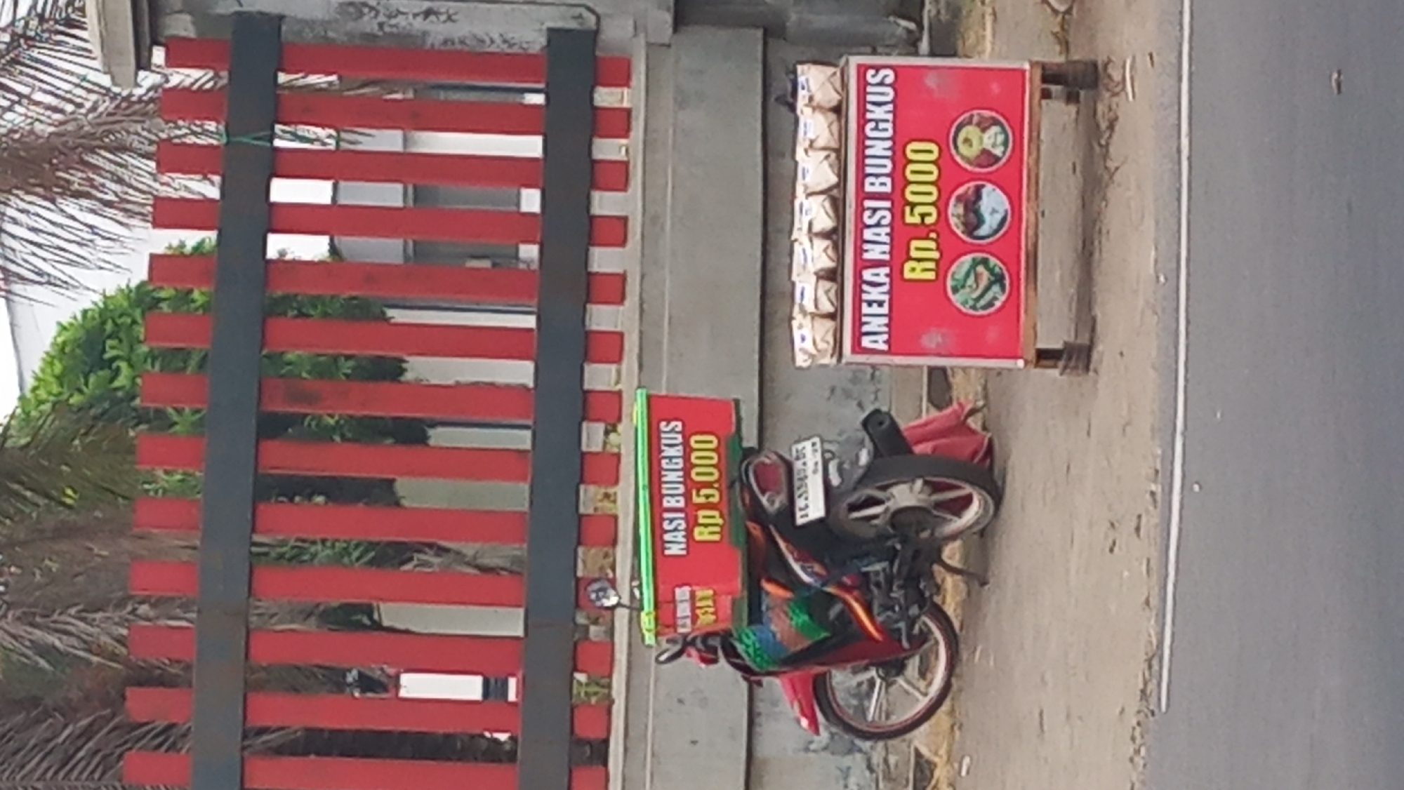 The image depicts a street scene in Pare, Indonesia, likely a roadside stall. A red and black striped fence is visible in the background. In the foreground, a red motorcycle is parked on the dirt ground. The motorcycle has a red food carrier box on its rear, labeled "NASI BUNGKUS Rp 5.000" in white and yellow text. To the right of the motorcycle, a red advertising board is propped up on wooden legs, displaying the text "ANEKA NASI BUNGKUS Rp. 5000" in white and yellow letters, accompanied by three circular images of food. Several individually wrapped food packages are lined up on top of the advertising board. A person's legs are visible beneath the motorcycle, wearing a red cloth. The overall environment suggests a commercial setting where food is being sold from a mobile unit.