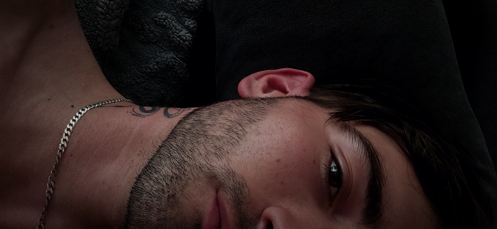 A close-up shot depicts a man lying down, angled with his left side slightly facing the camera. His face is partially visible, revealing a dark, trimmed beard and one eye. A silver chain necklace rests on his neck, and a tattoo is visible near his collarbone. His ear is also prominent. The background is primarily dark and out of focus. There are no indications of the city of Plovdiv, Bulgaria.