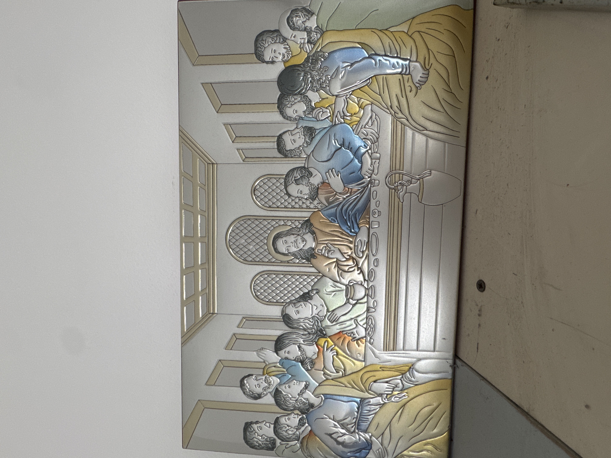 The image shows a metal bas-relief depicting the Last Supper. The scene includes Jesus and his twelve disciples seated at a table within a room with arched windows. The figures are rendered in a mix of silver, gold, and blue tones, with each disciple engaged in various expressions of emotion and interaction with Jesus. The bas-relief is mounted on a surface and appears to be located indoors, possibly in a church or religious setting. The exact location in the city of Varna, Bulgaria, cannot be ascertained from the image.