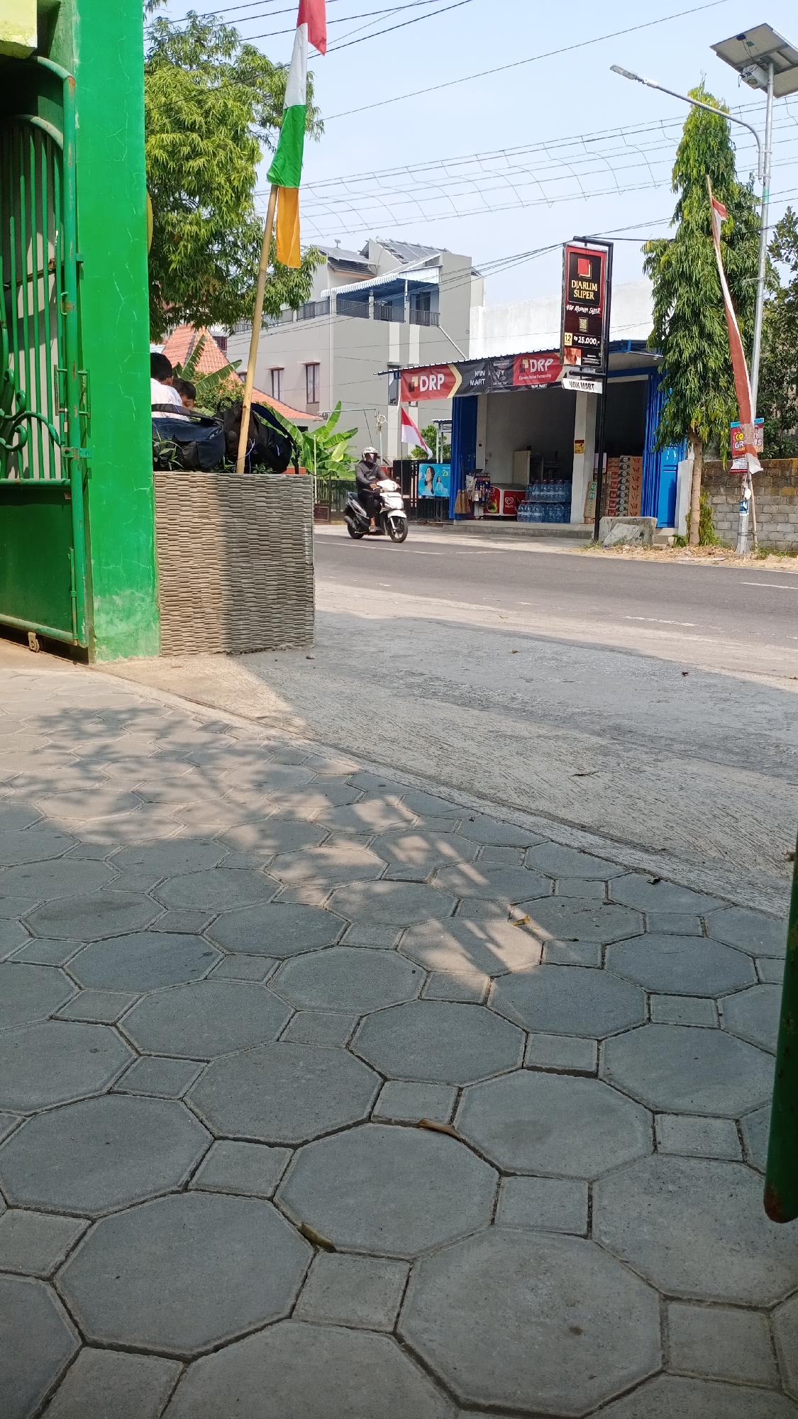 In Joho, Indonesia, a street scene unfolds under a clear sky. On the left, a vibrant green gate stands partially open, revealing a glimpse of a textured, light-colored wall. A flag with red, white, and green stripes flutters on a pole. A scooter rider, dressed in dark clothing and a helmet, is captured mid-motion on the asphalt road, heading towards the right. In the background, a row of buildings, including a shop with a blue facade and signage, lines the street. The ground in the foreground is paved with hexagonal and square concrete tiles, casting dappled shadows from unseen trees.