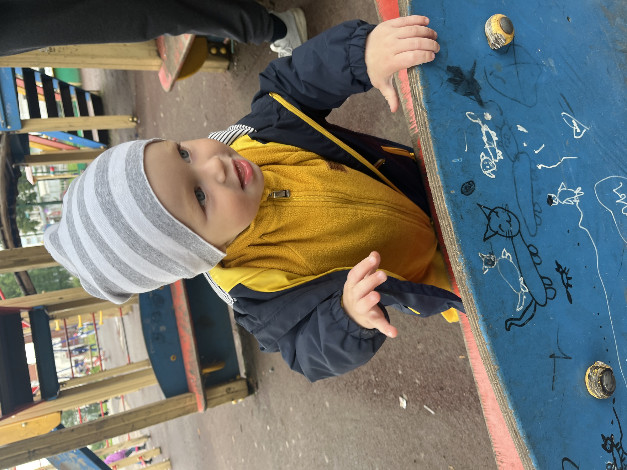 An outdoor close-up shot shows a young child, likely a toddler, wearing a grey and white striped beanie, a yellow fleece jacket, and a dark blue jacket with yellow trim, standing at a playground. The child is leaning on a blue and red playground structure, with their hands resting on the blue surface. The blue surface is decorated with various drawings, including a cat, a dog, and other figures. The background reveals a playground with wooden structures, swings, and other children playing. The ground appears to be a reddish-brown material, possibly rubber mulch or sand. The lighting suggests an overcast day. The location is identified as Chaikovsky, Russia.