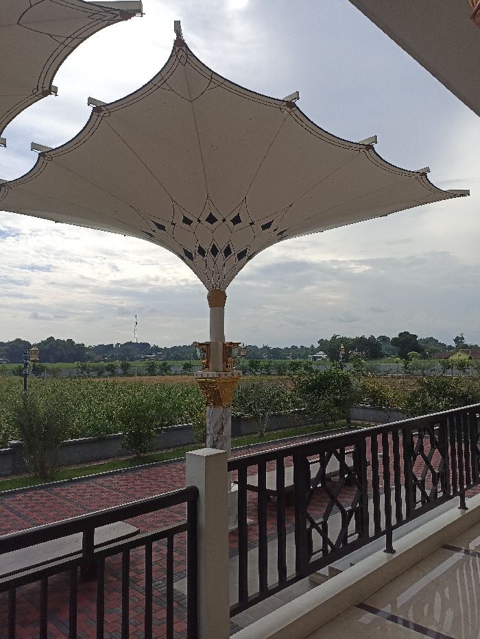 The image captures an outdoor scene dominated by a large, ornate, white tensile structure, resembling an umbrella or canopy, positioned centrally. Its fabric surface features intricate dark patterns, particularly around its central peak. This canopy is supported by a decorative pillar adorned with gold-colored accents and a greyish, marble-patterned base. Another similar canopy is partially visible on the upper left.

In the foreground, a black metal railing with geometric patterns suggests a balcony or an elevated viewing platform. Beyond the railing, a brick-paved path or area is visible.

The background unfolds into a wide agricultural landscape under a partly cloudy or overcast sky. Lush green trees and rows of plants, possibly crops or an orchard, stretch across the middle ground. In the distance, a tall, slender communication tower is visible, along with a small, white building topped with a pointed, possibly dome-like roof, likely a mosque or a similar religious structure, nestled among trees. The overall setting implies a blend of modern architectural features and natural, rural scenery in Plemahan, Indonesia, captured during daytime with soft, diffused lighting. No people or visible text are present in the image.