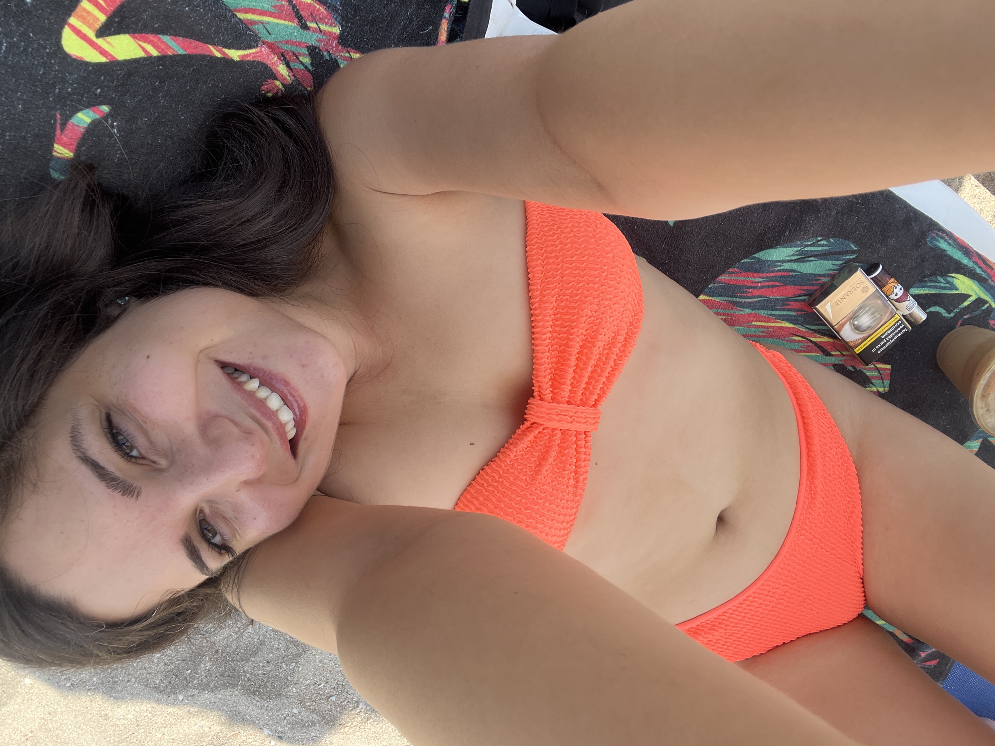 This is a first-person, overhead, close-up photograph of a young woman lying on a beach in Pomorie, Bulgaria. She is wearing an orange textured bikini and lying on a colorful beach towel or mat. Her head is tilted, and she is smiling at the camera, showing her teeth. She has dark brown hair that falls around her face and shoulders. The lighting suggests it is daytime, likely sunny, as indicated by the bright illumination and the shadows. In the upper right quadrant of the image, a pack of Sobranie cigarettes and a lighter are visible. The background shows sand and hints of the sea. The overall mood is relaxed and cheerful, suggesting a vacation or leisure activity.