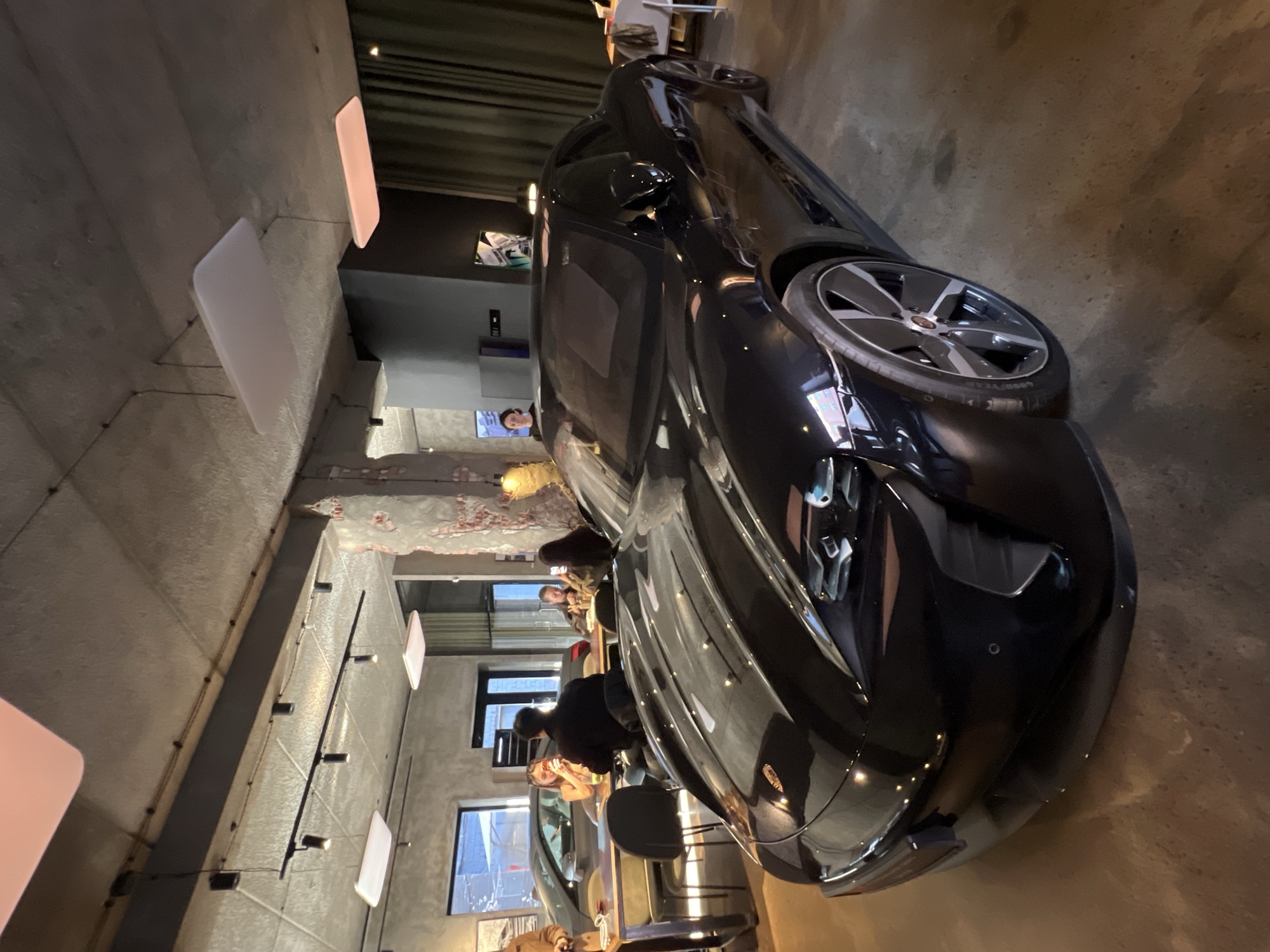 The image depicts a black car indoors, possibly a showroom or a restaurant in Saint Petersburg, Russia. People are seated at tables in the background. The car's front right tire is prominently displayed, showcasing its design and tread. The building's interior features exposed brick and modern lighting fixtures.