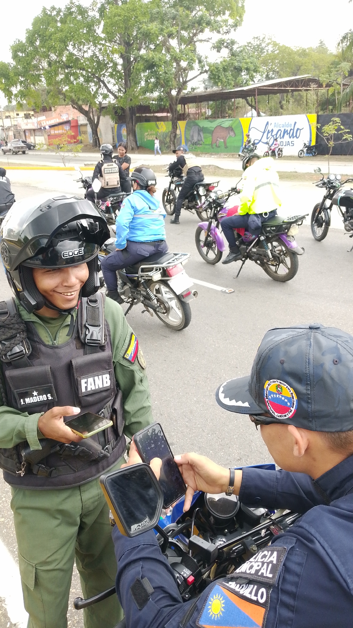In Tinaquillo, Venezuela, two uniformed officials interact on a street lined with parked motorcycles and a mural. One official, wearing a black helmet, green uniform, and a vest labeled "FANB," holds a smartphone and smiles towards another official in a blue uniform and cap emblazoned with the Venezuelan flag. The second official, also holding a smartphone, sits on a motorcycle with "LICIA MUNICIPAL TINAQUILLO" visible on their uniform. Behind them, several motorcyclists wait, some wearing helmets and light-colored jackets. In the background, a mural depicts wildlife, with the text "Fauna" and "Alcalde Lizardo" visible. A few pedestrians can be seen in the distance.
