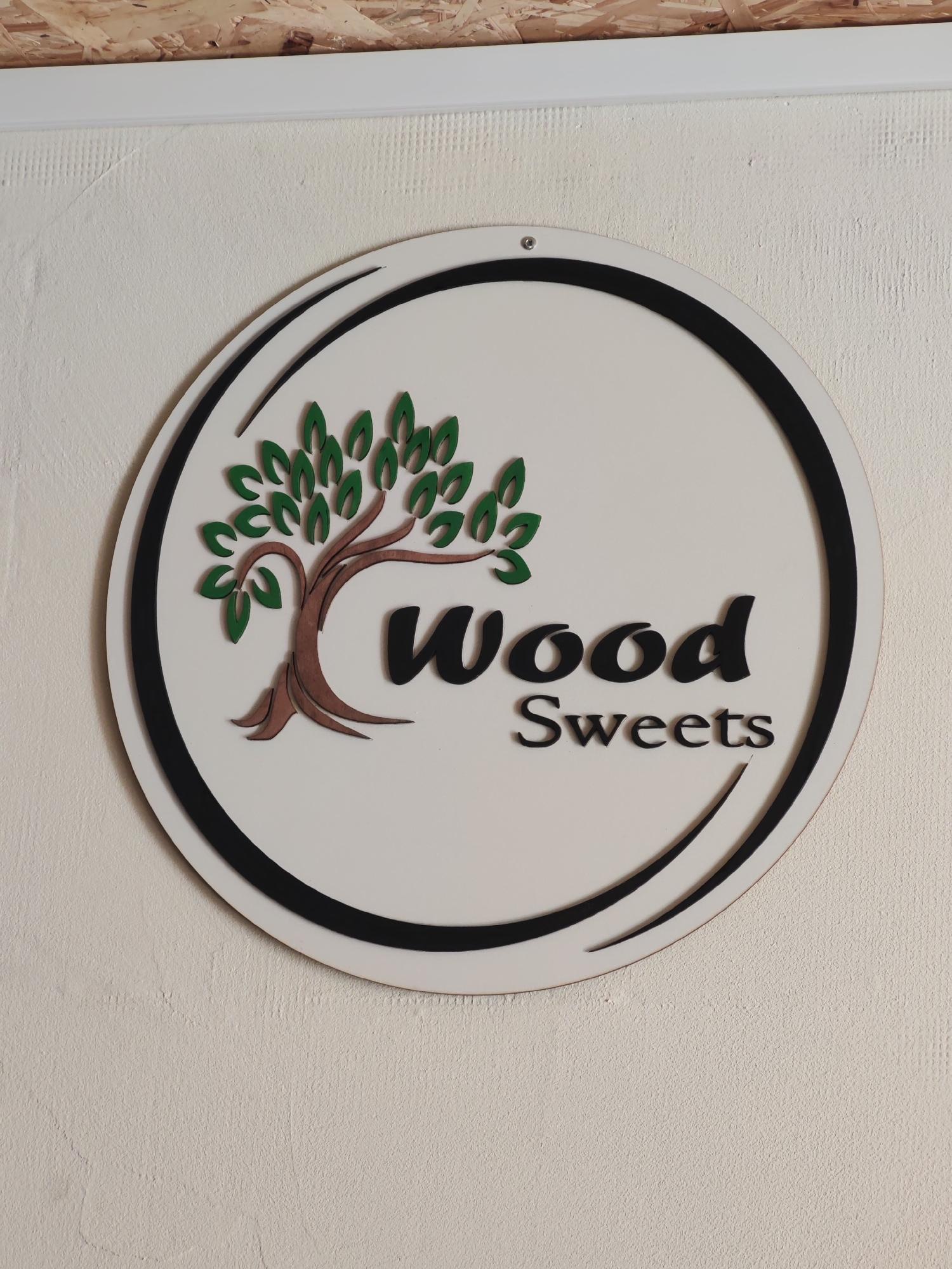 The image shows a round sign mounted on a textured white wall, likely indoors. The sign has a white background with a black outline and a stylized tree graphic on the left side. The tree has a brown trunk and green leaves. To the right of the tree, the words "Wood" and "Sweets" are written in a black, bold font. The location is Unknown, Unknown. There are no people visible in the image.