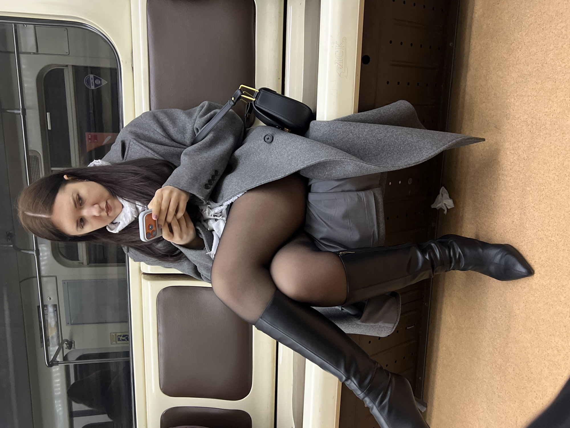 A woman is sitting on a train. She is wearing a gray coat, white shirt, black tights, and black knee-high boots. She is holding a cell phone. A black purse is on the seat next to her. The location is Shushary, Russia, given the carriage style. The train interior appears utilitarian.