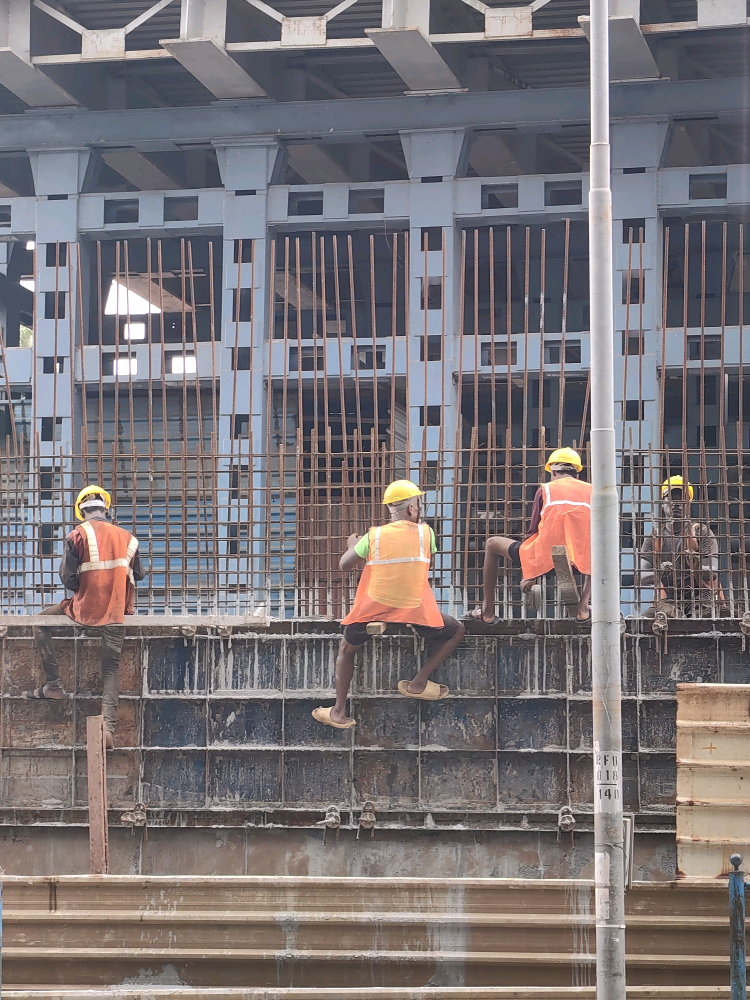 The image depicts a construction site in Mira Bhayandar, India. Four construction workers, all wearing yellow hard hats and orange reflective vests, are visible. Two workers are seated on scaffolding made of wooden planks, while the other two are actively climbing over a concrete formwork structure. The structure is reinforced with numerous vertical metal bars. In the background, there is a large, unfinished building with a metallic gray framework. A utility pole with some markings is also present to the right of the frame. The ground appears to be a mixture of concrete and debris.
