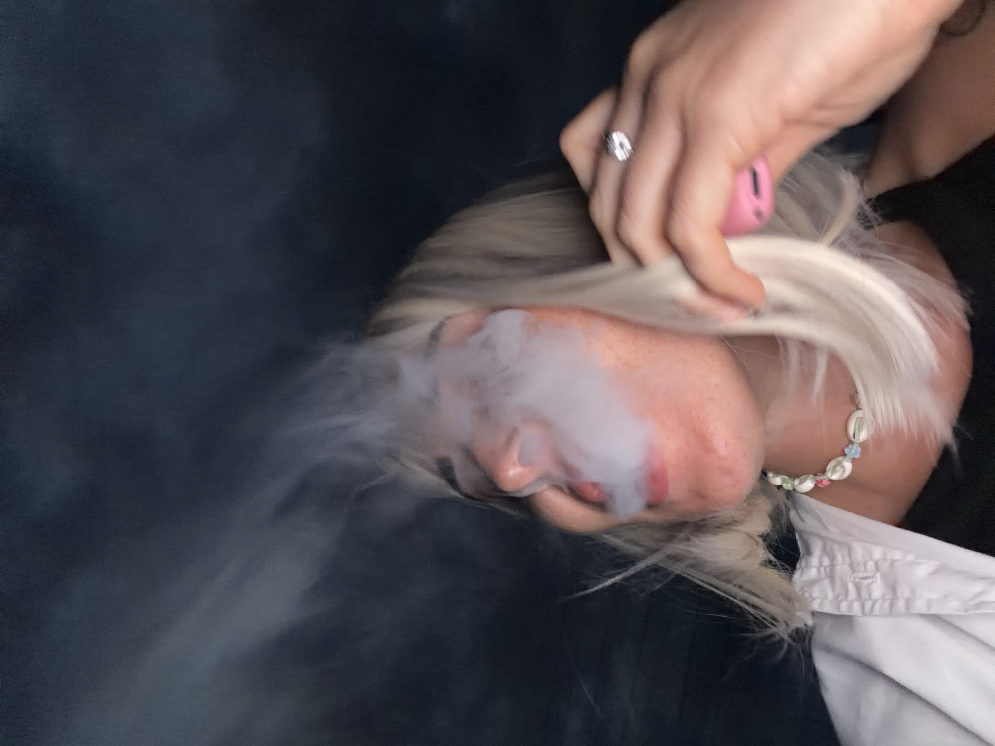 A close-up, medium shot captures a young woman with blonde hair exhaling a cloud of white vapor against a dark, blurry background. She is looking to her right, with her eyes partially closed, and her mouth open as the vapor escapes. The woman is wearing a white collared shirt over a dark top, a necklace made of shells and beads, and a diamond ring on her right ring finger. Her right hand is raised to her head, with her fingers brushing against her hair. A pink object, possibly a vape pen, is held between her thumb and index finger. The image appears to be taken at night or in a dimly lit environment. There is no identifiable information to suggest the location is in Sochi, Russia.