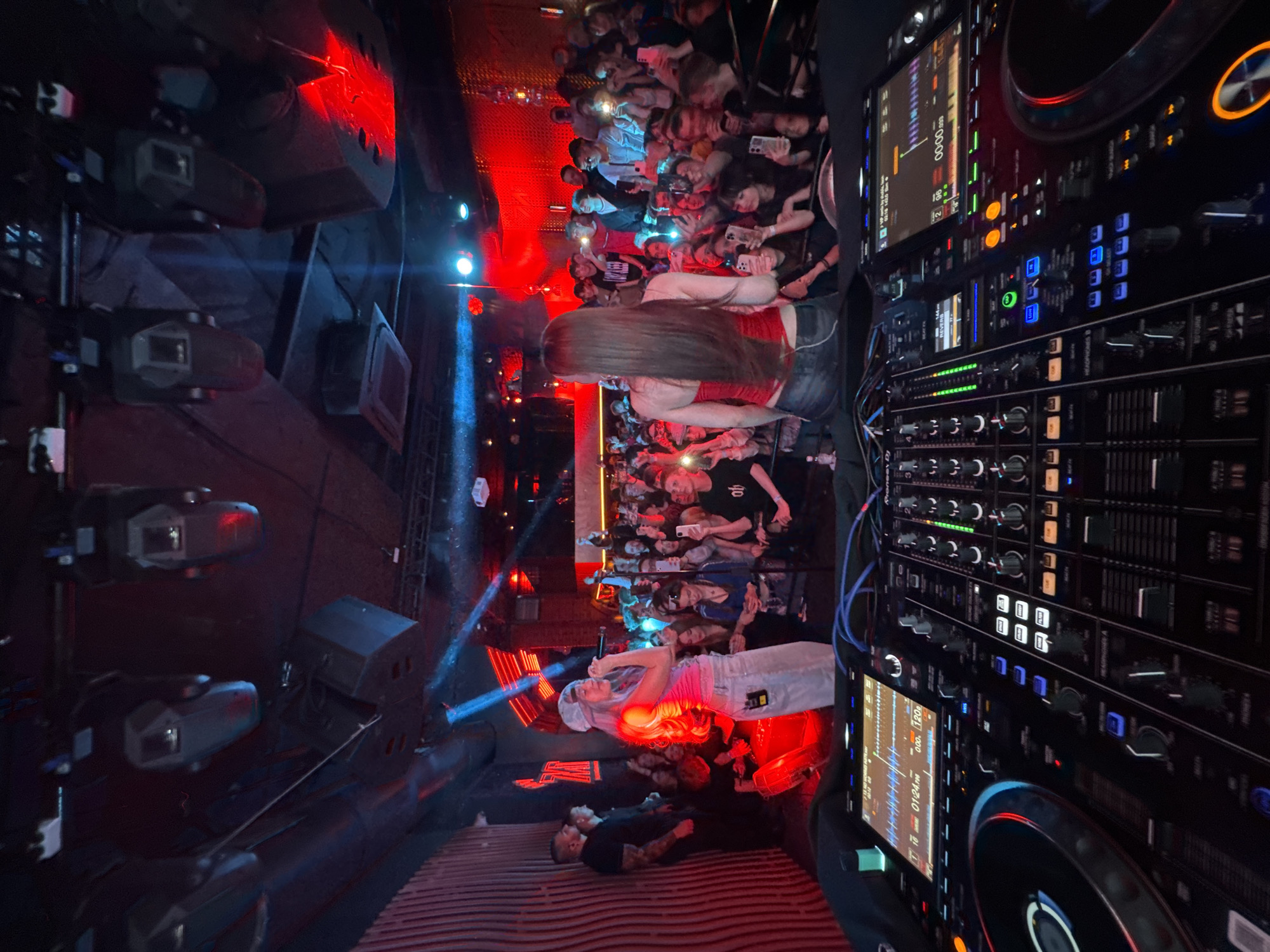 This image captures a vibrant nightclub or concert venue scene, filled with energy. The shot is taken from an elevated position, likely near a DJ booth, looking down at a stage and a crowded audience.
In the foreground, to the right, a substantial DJ setup is visible, featuring multiple Pioneer CDJs and a mixer with numerous buttons, faders, and illuminated screens displaying waveforms and track times, such as "00:00.033" and "01:24.700". Blue and red lights reflect off the equipment.
The central part of the image focuses on the stage and performers. A singer, dressed in a pink top and light-colored pants with a white head covering, holds a microphone and performs under bright red and blue stage lights. Another individual is seen lying or sitting on the stage floor further left. To the left of the stage, several large speakers or stage monitors are mounted, some with red lighting.
Beyond the stage, a dense crowd of audience members fills the floor. Many people are holding up their smartphones, presumably recording the performance or using their flashlights. A woman with long, straight dark hair, seen from behind, is prominent in the audience section closest to the stage. The overall atmosphere is dark, typical of a club, with dynamic lighting in shades of red and blue illuminating the performers and parts of the crowd. The visible text "Pioneer DJ" confirms the brand of the professional audio equipment. The event takes place in Khabarovsk, Russia.
