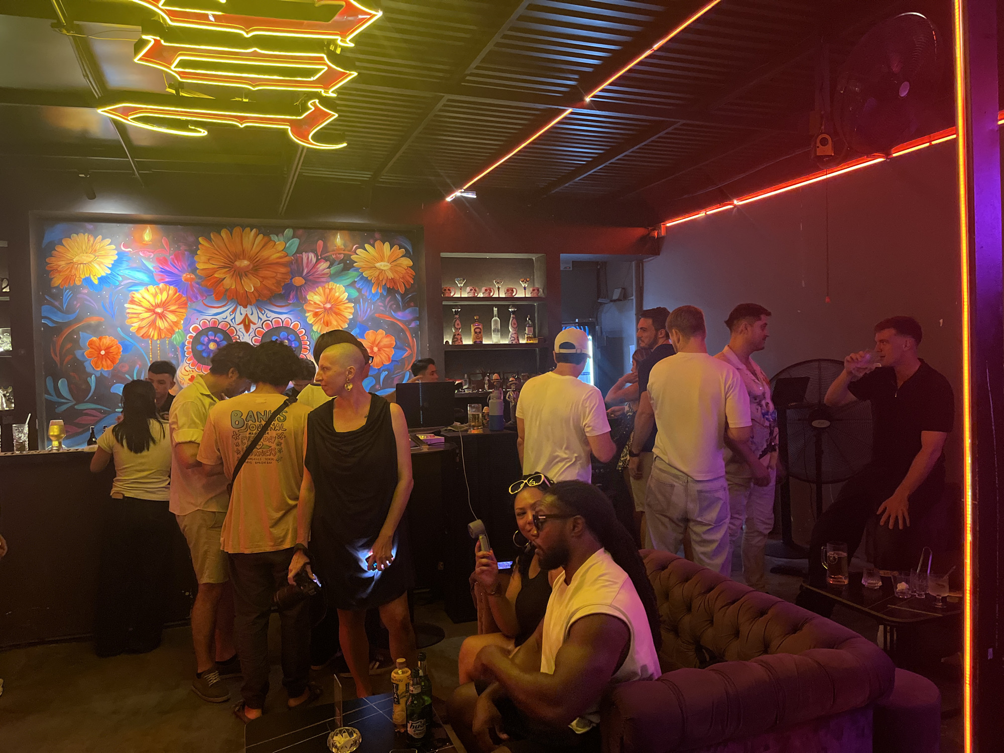 The image shows a crowded bar or lounge, likely in Đà Nẵng, Vietnam. A group of diverse patrons are mingling. Some are standing near the bar, while others are seated on a plush, tufted sofa. One individual is seated at a table drinking from a glass. A couple sits on the sofa, one holding a device, possibly a phone or remote. The interior has distinctive decor, including a mural with vibrant floral designs and skull motifs, likely inspired by Día de Muertos. Neon lighting fixtures are mounted on the ceiling, with both linear orange lights and decorative yellow arrows. The overall lighting is dim, contributing to a lively, evening atmosphere. Bottles are visible on shelves behind the bar. A large industrial fan is mounted on the wall in the background.