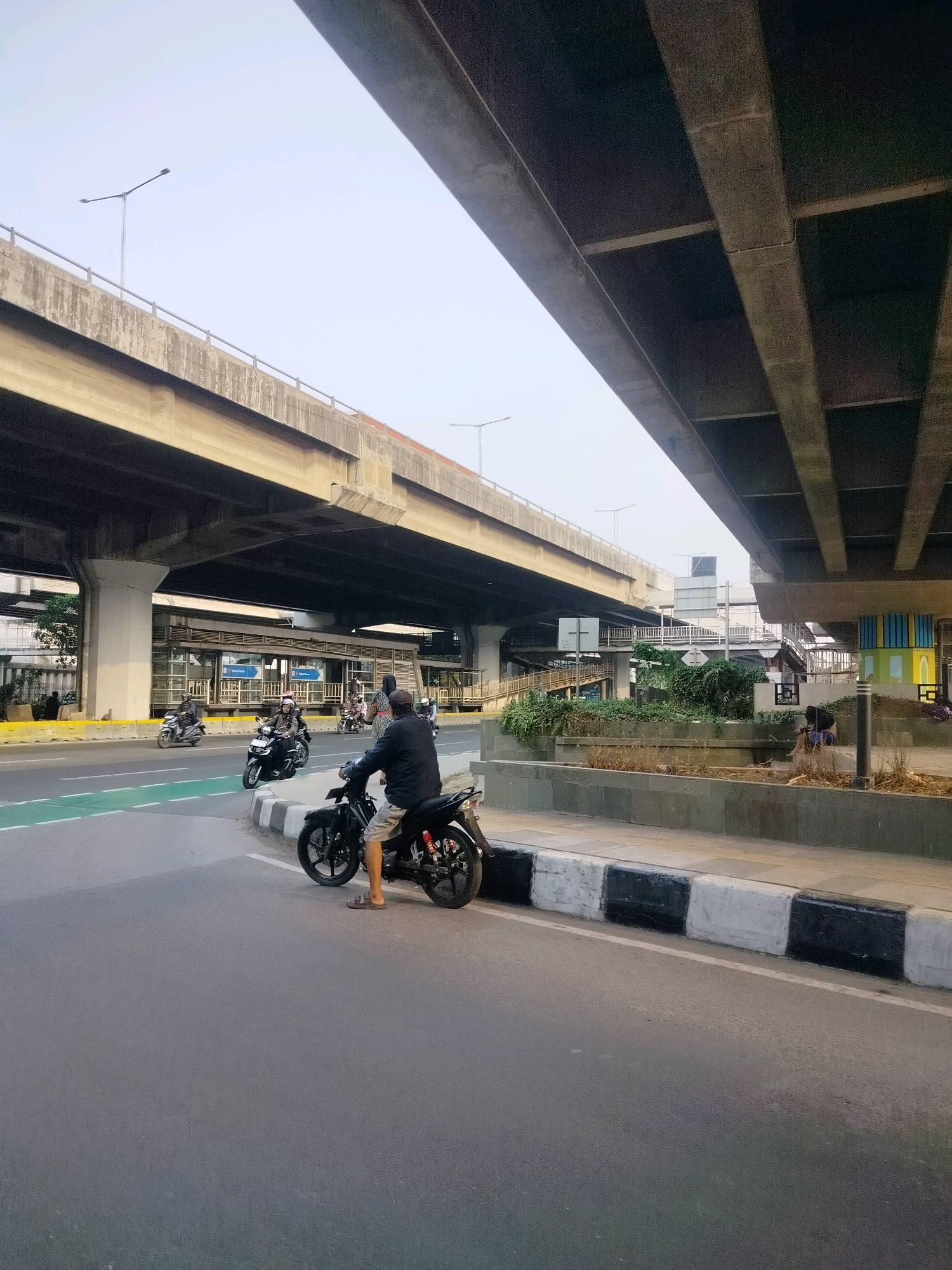 This is an eye-level shot of a street scene in East Jakarta, Indonesia, under a massive elevated highway. In the foreground, a person on a black motorcycle is stopped on the right side of the road, facing left, with their legs extended. They are wearing a dark jacket and light-colored shorts. Behind them, other motorcycles are visible on the road, including one with a passenger wearing a helmet. The road has a green bike lane on the left. The scene is dominated by the massive concrete structure of the elevated highway, with its layered beams and supports. A pedestrian walkway with a black and white striped curb runs alongside the road. In the background, there are buildings, more elevated roads, and greenery. The sky is overcast.