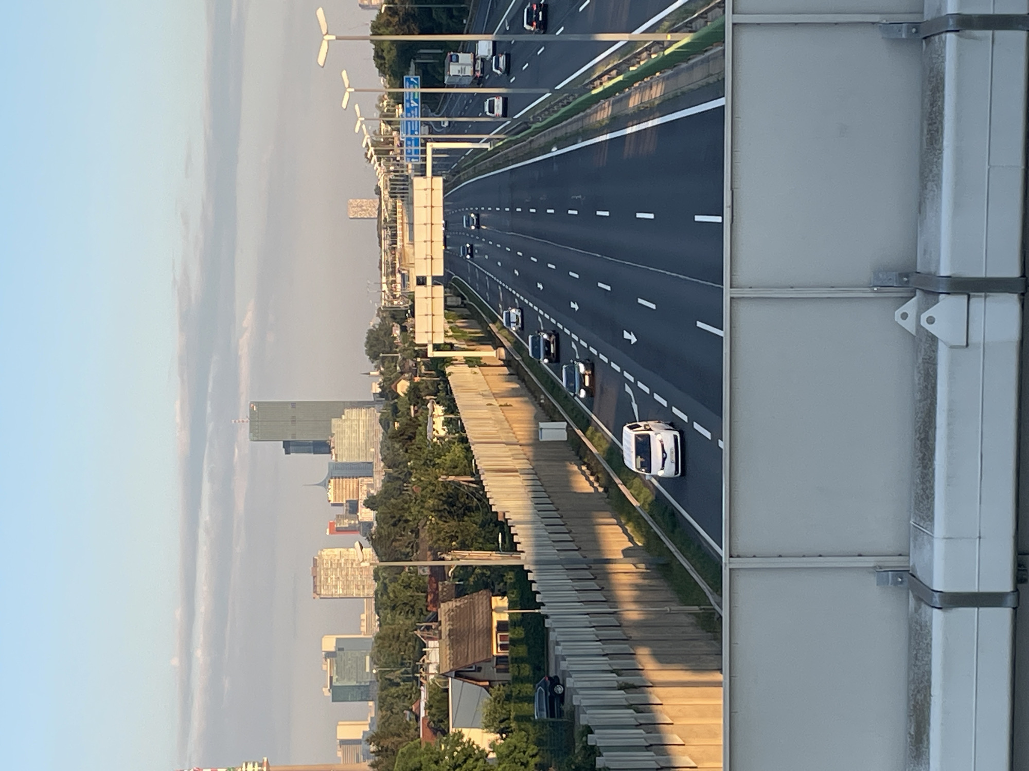 The image captures a view of a multi-lane highway stretching into the distance, with a cityscape visible on the left side. The highway is busy with several cars driving in the same direction. The time of day appears to be late afternoon or early evening, as indicated by the long shadows cast by the highway barriers and the warm, soft lighting. The sky is a clear blue with some wispy clouds.
On the left side of the highway, a dense urban landscape is present, featuring numerous tall buildings, suggesting a major city. The architectural styles of the buildings vary, with some appearing to be modern skyscrapers and others older structures. Green trees are interspersed throughout the cityscape and along the edge of the highway.
The highway itself is well-maintained, with clear lane markings. A sound barrier runs along the right side of the highway, partially obscuring the view of the immediate surroundings. A few wind turbines are visible in the distance on the left side of the highway, near the cityscape, and what appear to be streetlights or lampposts line the highway's edge.
No people are clearly visible as distinct subjects, but the presence of cars implies their activity of commuting or traveling. The overall mood is one of urban dynamism and movement.
There is no visible text in the image that can be read or interpreted. The location context is Vienna, Austria, which aligns with the modern urban architecture and the presence of wind turbines.