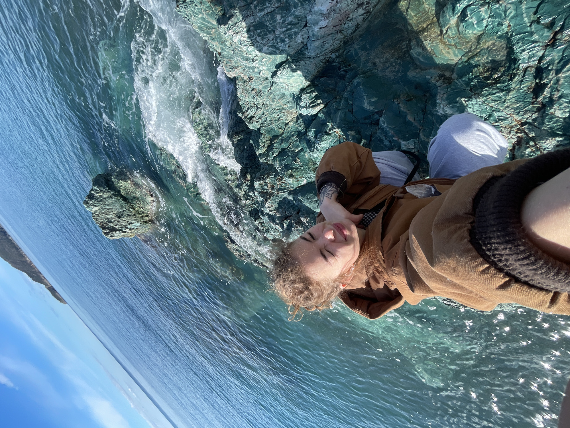A photograph taken at an angle depicts a person lying on blue-green rocks and turquoise water, likely by a waterfall. The person has fair skin and curly blonde hair, wearing a brown jacket and light-colored pants. The water is clear with sun glare on its surface. In the distance, the sea meets a light blue sky on the horizon. The photo appears to be taken from a low angle. The location is Rafailovići, Montenegro, evidenced by the rocky coastline and water color.