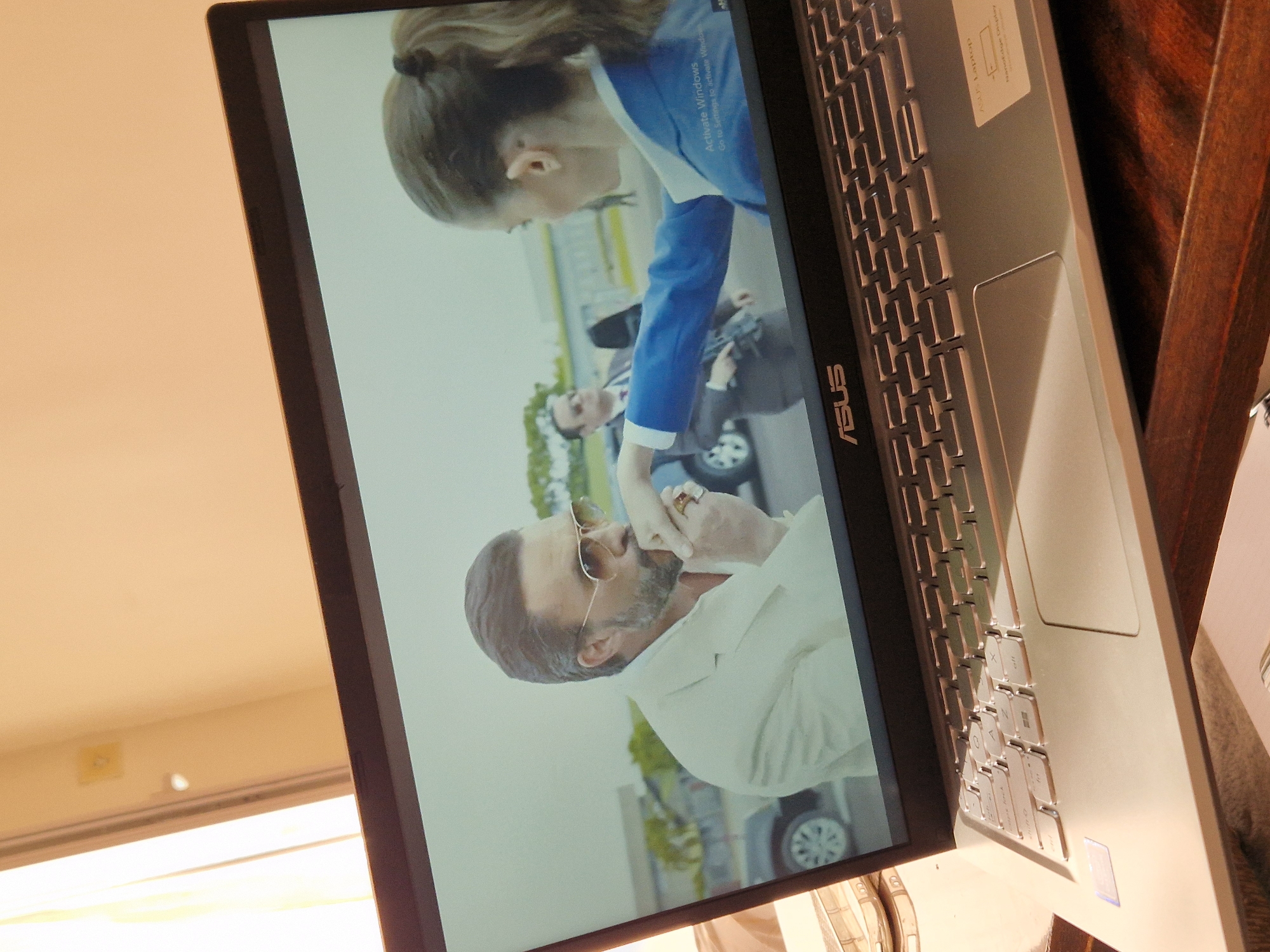 The image shows a laptop screen displaying a scene with two people. On the left, a man with sunglasses, a beard, and a white suit is kissing a woman's hand. The woman, on the right, is wearing a blue jacket and her back is to the camera. In the background, there are cars and some greenery. The laptop is open, resting on a wooden surface, with a keyboard visible in the foreground. The image does not provide any information about the location being Lovech, Bulgaria.