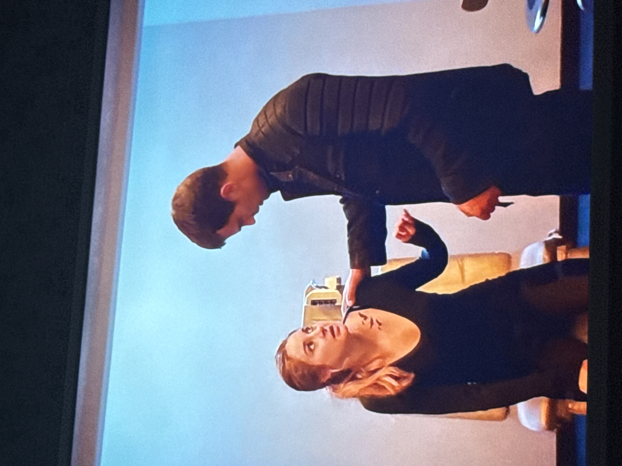 A man in a dark jacket leans over a woman in a dark outfit. They are indoors with a light blue wall and furniture in the background. The location of the image is unknown, and there is no way to tell if this is in Mytishchi, Russia.