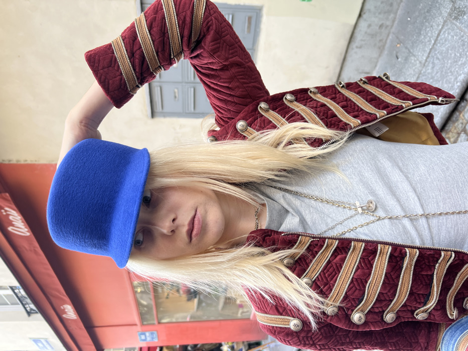A blonde individual is depicted in a close-up, outdoors on a city street in Paris, France. They wear a vivid blue felted hat with a short brim, which they are holding with their right hand placed on the hat's crown. Their long, light blonde hair frames their face, and they look towards the viewer's left. They are dressed in a dark red (maroon) quilted jacket with a prominent military-style design featuring multiple horizontal bands of golden-tan braided trim secured by silver-toned buttons, some marked "HP." The jacket is unzipped, revealing a light grey cowl-neck shirt and several silver-toned necklaces, including a choker and a longer chain with a clear, possibly glass, pendant. In the background, a building with a light-colored facade is visible. A shopfront with a bright red awning or facade element displays the name "Anaïs" in white script. A partial blue street sign is mounted on the building to the left of the shop. Further right, against a lighter wall, are several grey rectangular utility boxes or mail slots. The street paving consists of light-colored blocks.